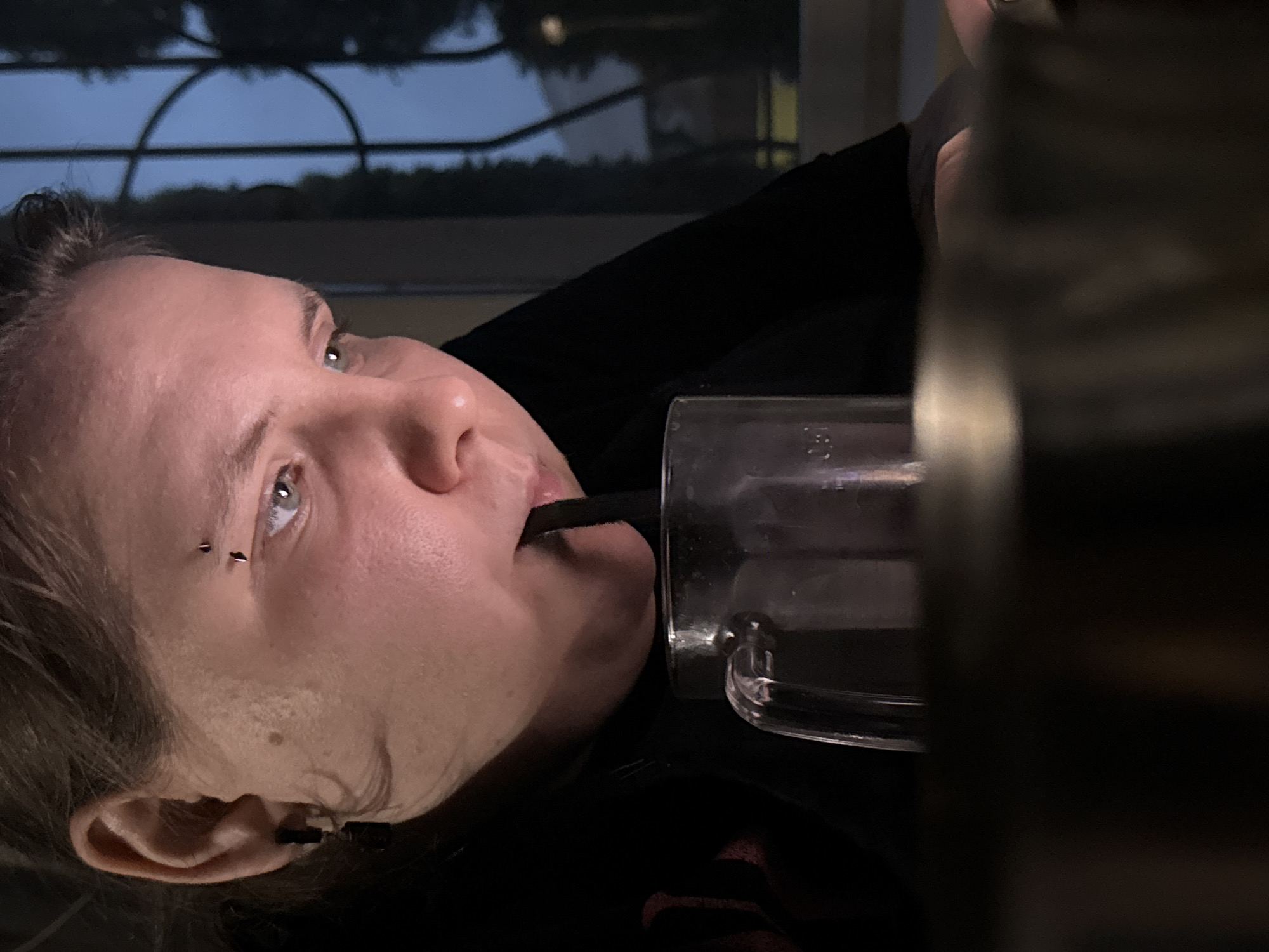 An extreme close-up shot shows a person drinking from a clear glass mug with a dark straw. The person's face is partially obscured by the mug and straw, but their light-colored eyes and eyebrow piercing are visible. Their head is tilted downwards, and they appear to be looking at something below the frame. The background is dark and blurry, with faint hints of trees and window panes, suggesting an indoor setting at night. The lighting is dim, casting a warm glow on the person's skin. The specific location in the city of Zaokskiy, Russia is not discernible from the image.