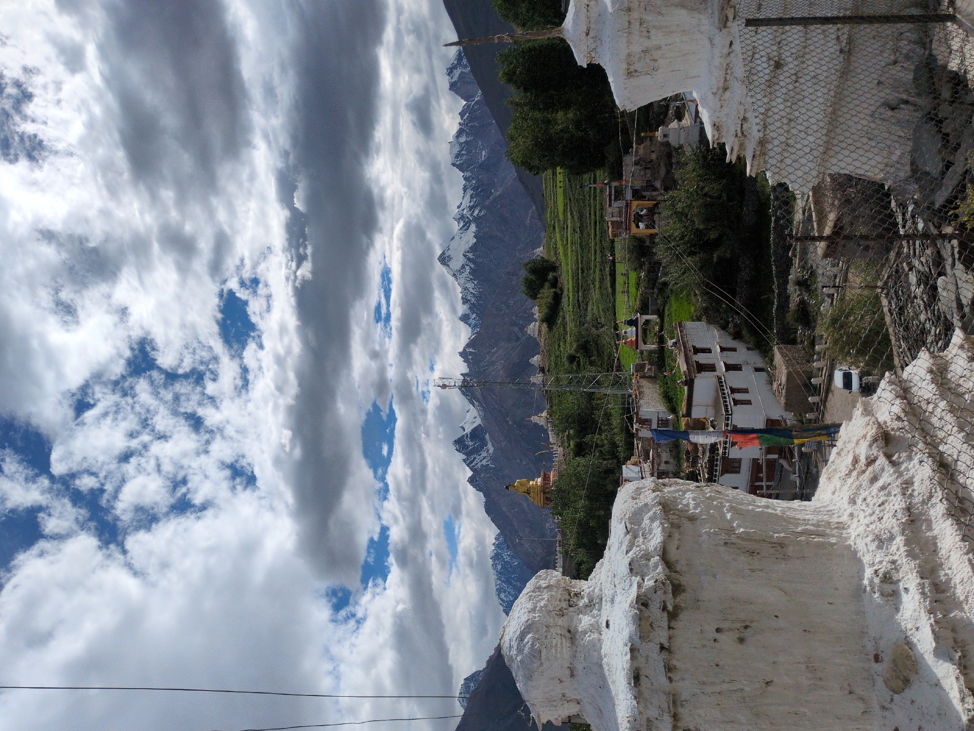 This is a photograph of the Karsha Monastery in Karsha, India, with a view of the surrounding landscape. In the foreground, to the left and right, are two whitewashed stupas, traditional Buddhist reliquaries. The stupa on the left is partially visible, showing its textured, weathered surface. The stupa on the right is more fully in view, with a chain-link fence in front of it.
In the middle ground, a village with white buildings and flat roofs is nestled in a green valley. Prayer flags in vibrant colors of red, blue, yellow, and green are strung between some of the buildings. Behind the village, a large, golden Buddha statue sits on a pedestal, overlooking the scene. Beyond the village and the Buddha statue, terraced fields climb the hillsides, dotted with trees.
In the background, a majestic mountain range with snow-capped peaks rises against a dramatic sky filled with white and grey clouds. A telecommunications tower is visible on a ridge to the right of the Buddha statue. A white car is parked on a road near the village. The overall impression is one of a remote, spiritual location set against a stunning natural backdrop.