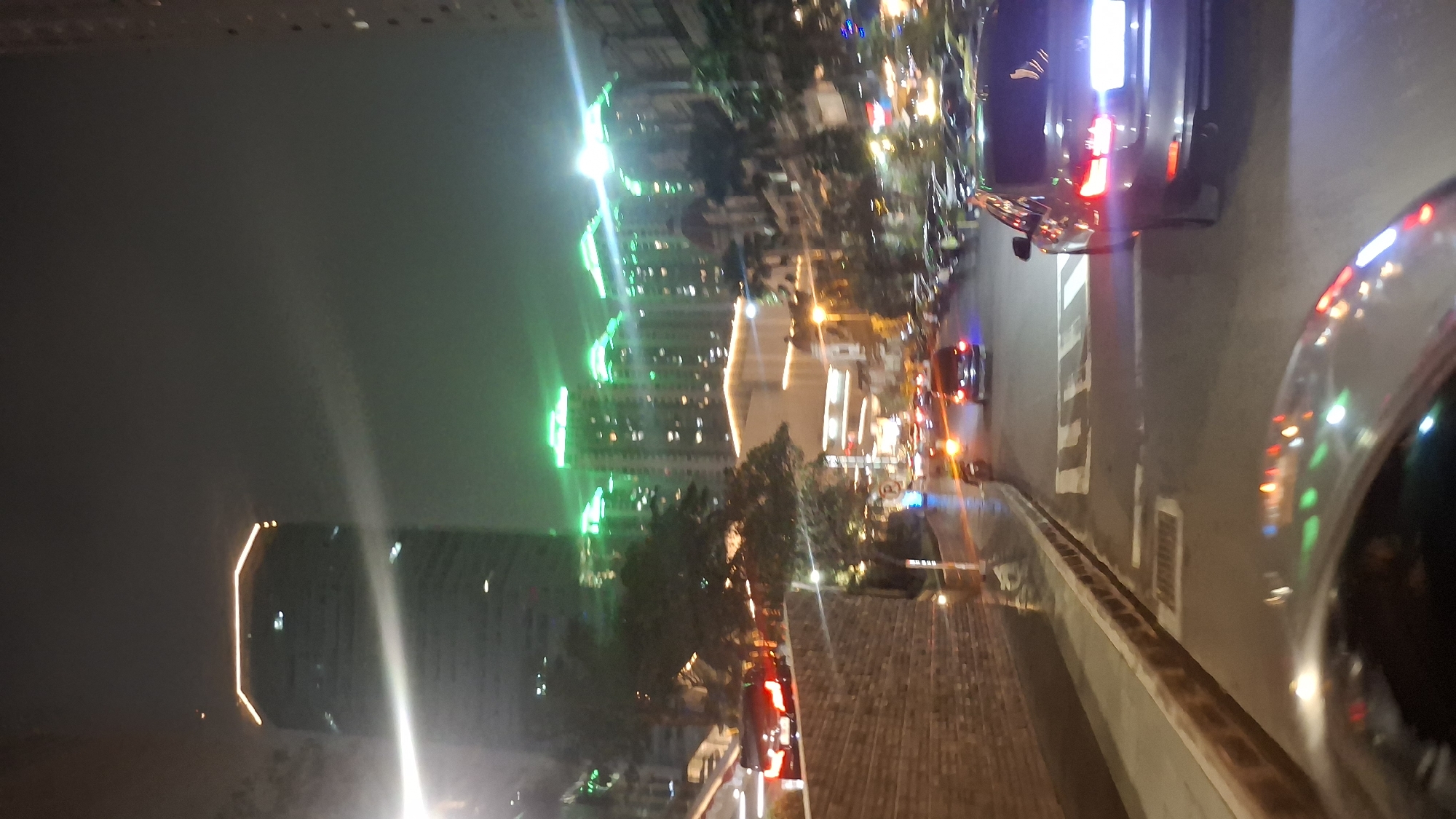 The image captures a nighttime scene in a city, possibly West Jakarta, Indonesia. A tall building with illuminated orange lights on its upper edges dominates the left side of the frame. To the right, a series of high-rise buildings are lit with vibrant green lights along their rooftops. The foreground shows a street with several cars, including a dark-colored sedan with its brake lights illuminated, suggesting it is either stopped or slowing down. The road surface appears wet, reflecting the colorful lights from the buildings and vehicles. Trees line the street, and brickwork is visible on the left side of the image. The overall atmosphere is one of an urban environment at night, with abundant artificial lighting.