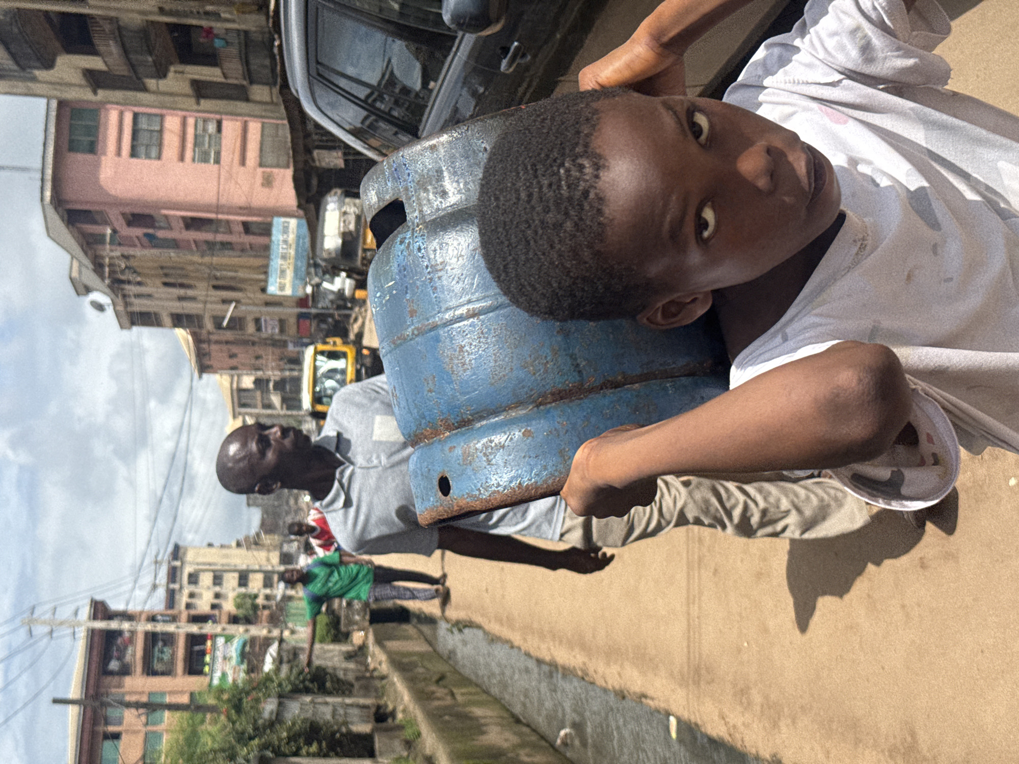 In Onitsha, Nigeria, a young boy, center frame, carries a large, blue, cylindrical object on his shoulders, partially obscuring his face. He wears a white t-shirt with a patterned design. Behind him, an adult man in a light grey polo shirt and beige pants walks away from the camera, also carrying the same blue object. In the background, several people are visible, some walking, others standing. The scene takes place on a dirt path alongside a narrow, murky canal. Buildings of varying heights and colors line the path, suggesting a dense urban environment. Vehicles, including a yellow truck and a black SUV, are present in the background. The sky is partly cloudy.