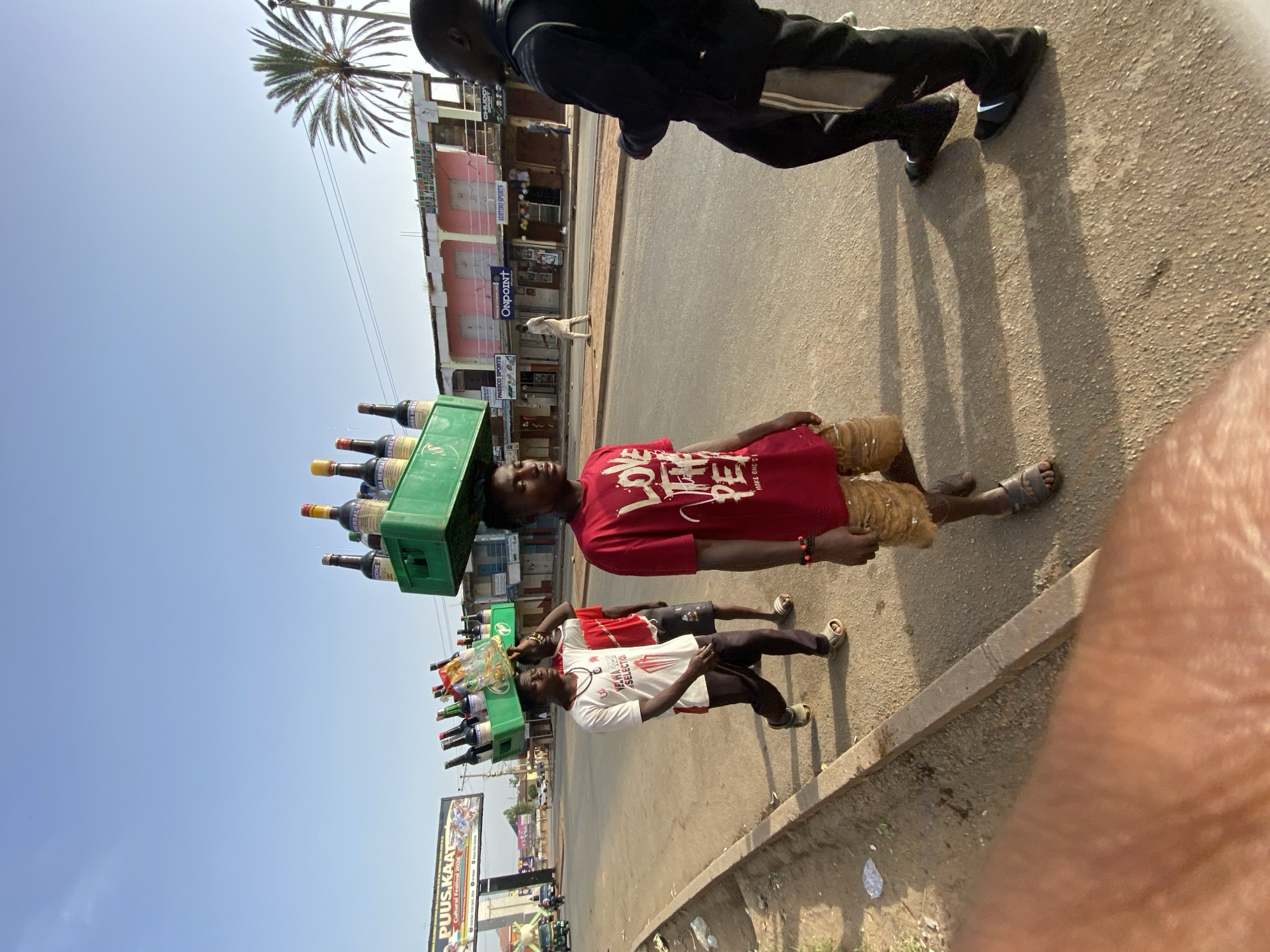 The image depicts a street scene in Jos, Nigeria. A man is carrying a green crate full of bottled drinks on his head, walking along the sidewalk. He is wearing a red t-shirt and tan shorts. Another man is walking in front of him, also carrying a crate on his head, wearing a white shirt and dark pants. There are shops and buildings in the background. A billboard on the left side reads "PUUS KAAT Cultural Festival 2026". The street is paved and lined with a curb. Another man in jogging pants walks in the opposite direction.