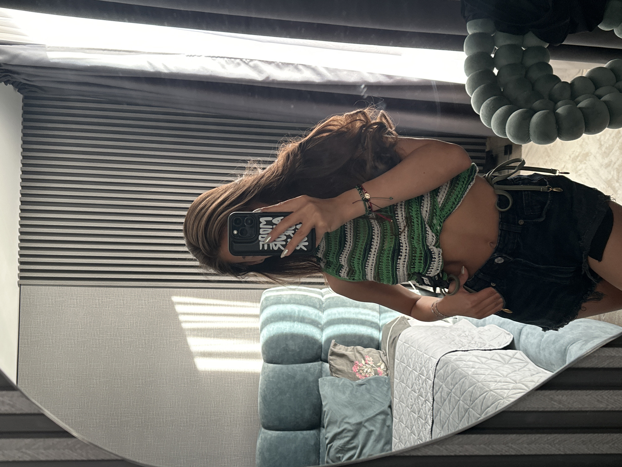 A young woman with long, brown hair is taking a mirror selfie in a bedroom. She is wearing a green and white striped crochet halter top and black denim shorts. She is holding a smartphone in her right hand, which is angled to capture the reflection. The bedroom has a large, oval mirror on the left, a bed with a quilted gray comforter and blue pillows on the left, and vertical striped wallpaper and grey curtains on the right. The lighting suggests it is daytime, with sunlight streaming through the curtains. The overall impression is of a casual, personal moment. The location is specified as Sofia, Bulgaria.