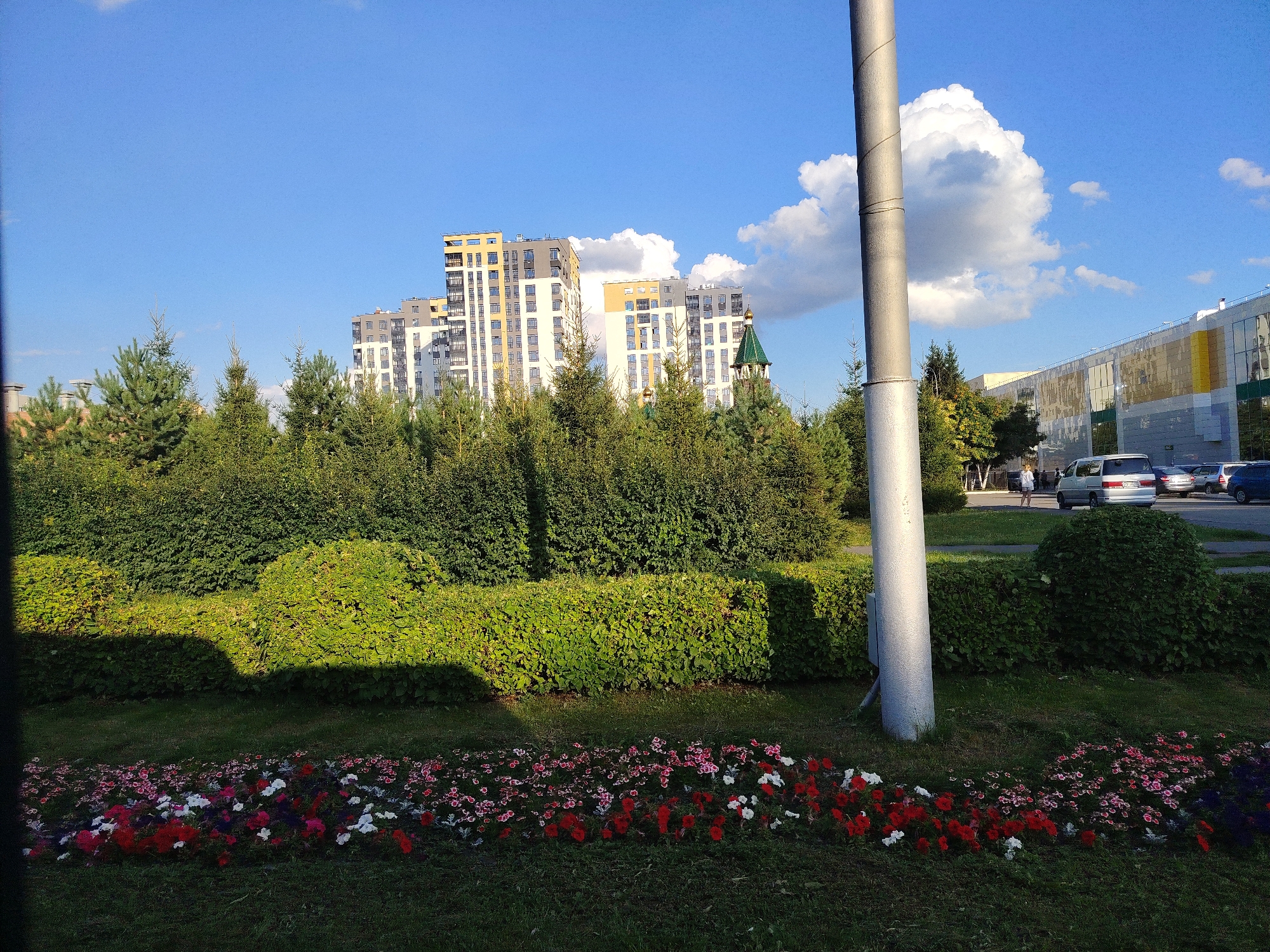 The image shows a well-maintained urban park in Kemerovo, Russia, featuring a foreground of colorful flowerbeds with red, white, and pink blossoms arranged in neat rows. Behind the flowers, manicured green bushes create a layered landscape. In the middle ground, several evergreen trees, likely pines, stand tall amidst the foliage. The background reveals a modern cityscape with tall apartment buildings exhibiting a mix of white, gray, and yellow facades, punctuated by a green-domed tower, possibly part of a church. To the right, a large, light-colored building with prominent windows and yellow accents appears to be a commercial or public facility. A light pole stands prominently in the right foreground, casting a shadow on the bushes. A street is visible in the distance with several cars parked and a few people walking, suggesting a bustling urban environment. The sky is a clear blue, with a large, fluffy white cloud dominating the upper right portion of the frame, indicating fair weather. The overall scene depicts a blend of nature and urban development.