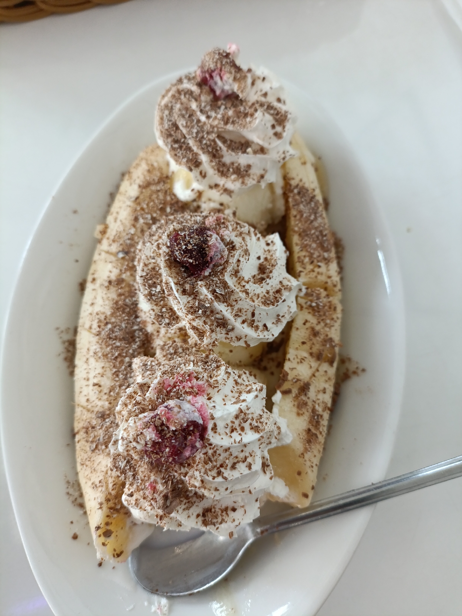 An overhead shot shows a banana split dessert on a white oval plate. The dessert consists of a banana cut lengthwise, topped with three dollops of whipped cream. Each dollop of whipped cream is sprinkled with chocolate shavings and a small amount of red berry topping. The banana itself is also dusted with chocolate shavings. A silver spoon rests on the right side of the plate, partially submerged in the dessert. The background is a white surface, likely a table, with the corner of a woven basket visible in the upper left. There is no discernible location in Chaikovsky, Russia, or any people present in the image.