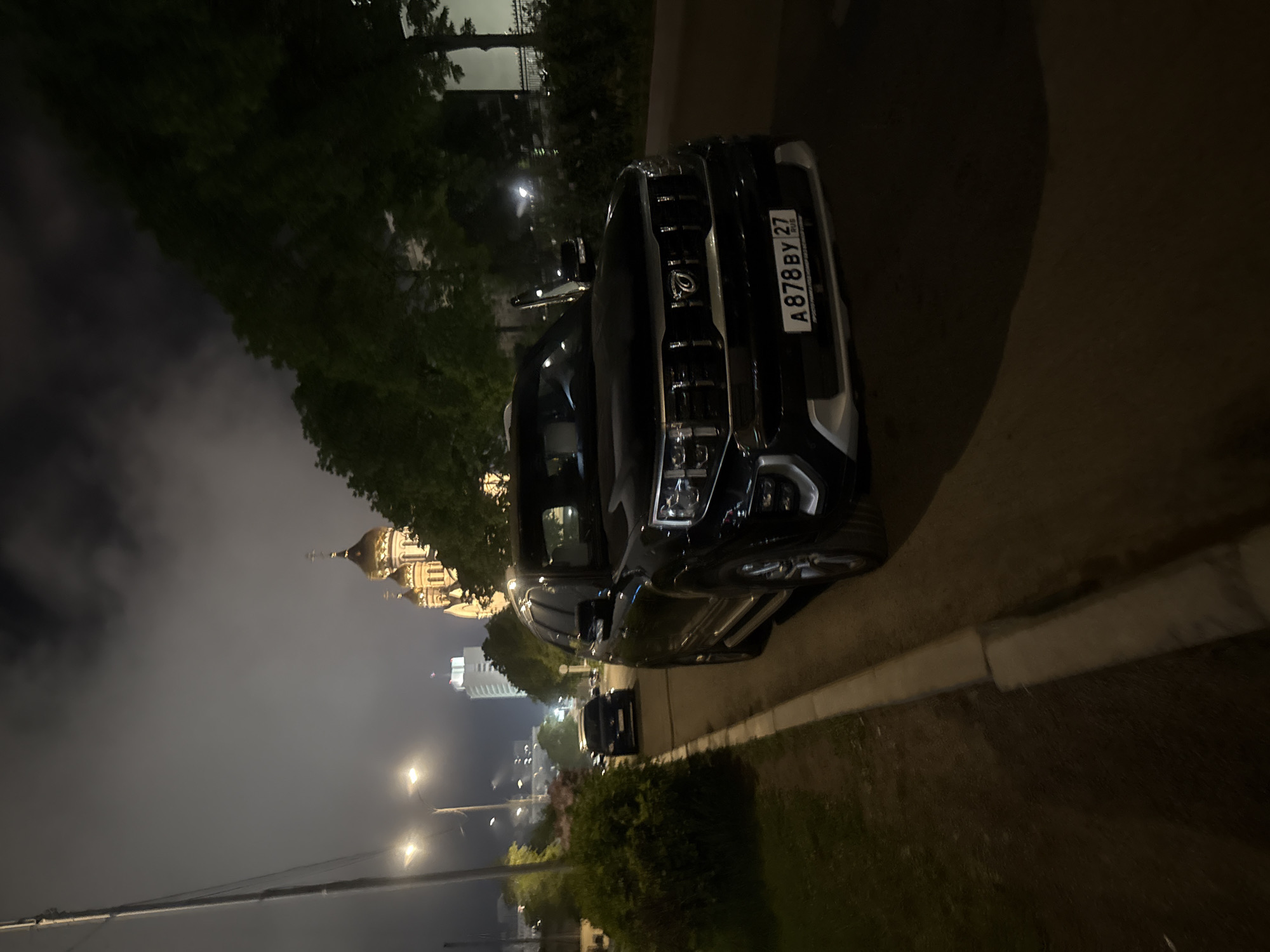 The image depicts a nighttime street scene in Vladivostok, Russia. A black SUV is parked on the right side of the road, its front facing towards the viewer. The SUV has its license plate visible, reading "A878BY 27 RUS". In the background, a distinct golden-domed church with multiple spires is visible, along with a tall, modern white building. Trees line the right side of the road behind the SUV, and a streetlight illuminates the left side of the street. A smaller dark-colored car is parked further down the road on the left. The sky is dark and cloudy, with some atmospheric haze. The scene appears to be a quiet urban environment at night.