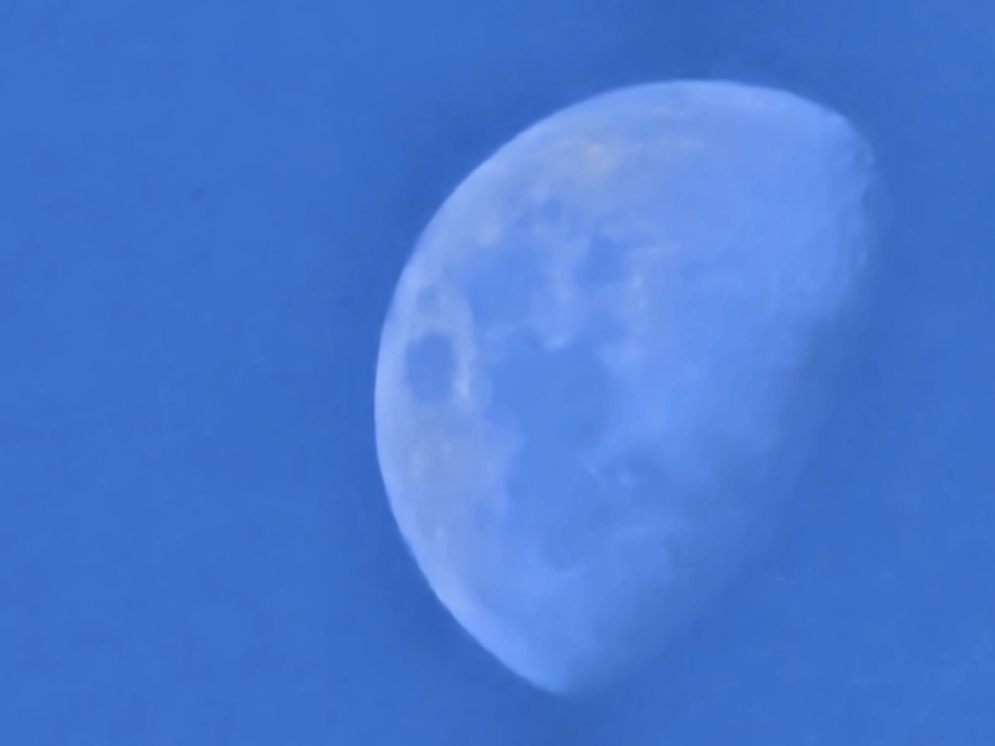 An image taken in Esch-sur-Alzette, Luxembourg, depicts a partially illuminated celestial body, identified as the Moon, against a clear, uniform blue sky. The Moon occupies the lower-middle portion of the vertical frame, with approximately three-quarters of its disc visibly illuminated in a pale, almost milky blue tone. The illuminated edge is on the right side of the visible disc. Surface features on the Moon are discernible but appear softened, possibly due to atmospheric conditions or focus. The background sky is a consistent, vibrant medium blue, indicating daytime or twilight conditions. No other objects, subjects, or actions are present within the frame.