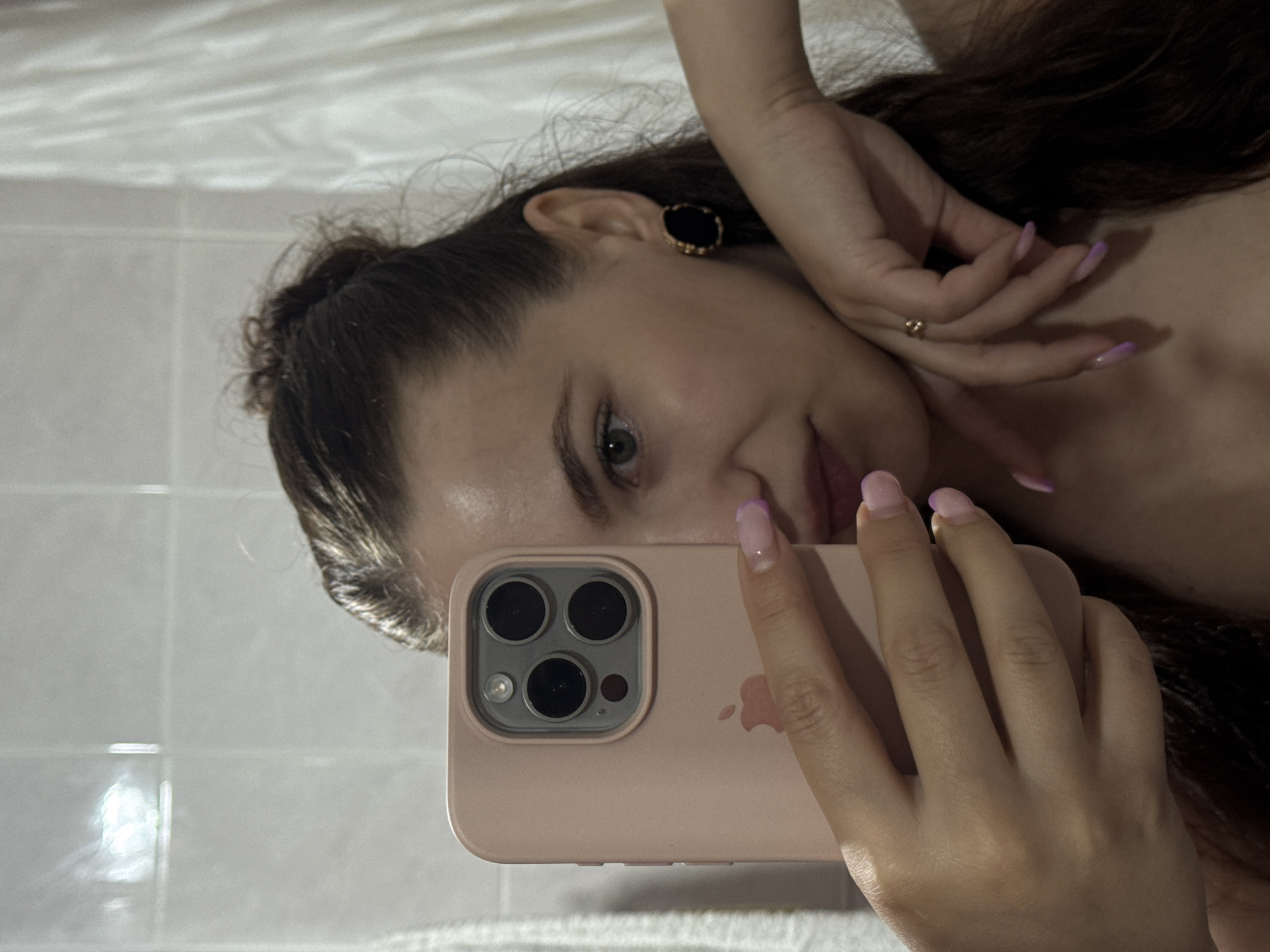 The image is a mirror selfie of a young adult female in an indoor setting. The subject's face is partially visible, with her right eye, nose, and mouth showing a neutral to slight smirk expression. Her left cheek and the majority of her face are obscured by a pink/peach-colored iPhone, held in her left hand, displaying its triple-lens camera array and partially visible Apple logo. Her dark brown hair is pulled back, possibly in a bun, with some strands loose around her face and shoulder.
The subject's right hand rests on her upper chest or left forearm, with fingers gently splayed. Both hands feature manicured nails with a light purple/pink ombré or French tip design. A small, gold-toned ring is visible on the ring finger of her right hand. On her right earlobe, a black, circular earring with a gold-toned rim is present. The subject appears bare-shouldered, revealing her décolletage.
The background consists of white tiled walls, characteristic of a bathroom, with visible grout lines and a bright light reflection on the upper tiles. To the far right, a white shower curtain or hanging towel is visible. There are no discernible visual cues or objects within the image that specifically indicate the city of Chaikovsky, Russia.