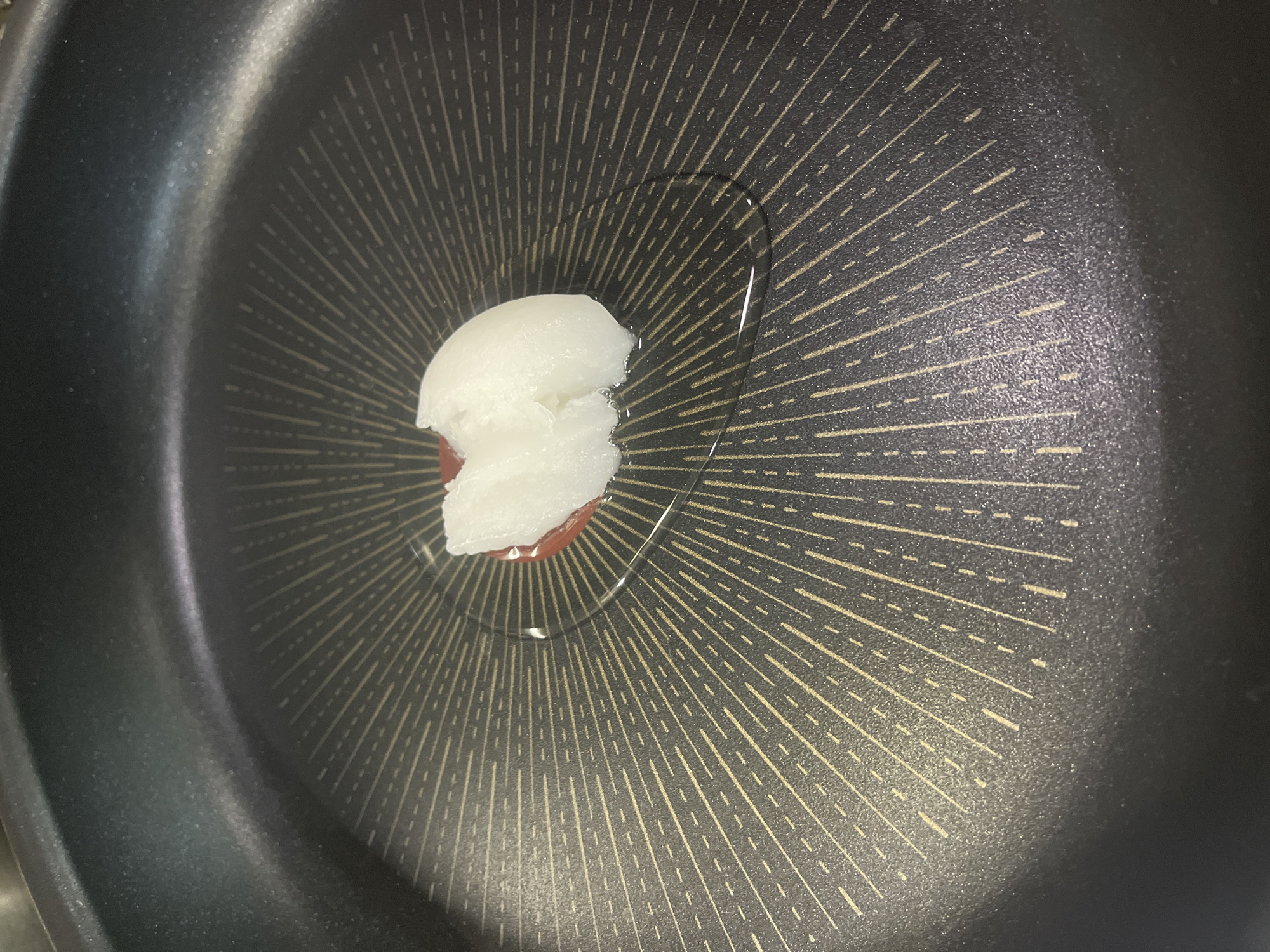 A close-up shot shows a black non-stick frying pan with a piece of solid white coconut oil melting in the center. A small amount of clear liquid cooking oil surrounds the coconut oil. The pan's surface has a pattern of radiating lines. The location is not identifiable from the image.