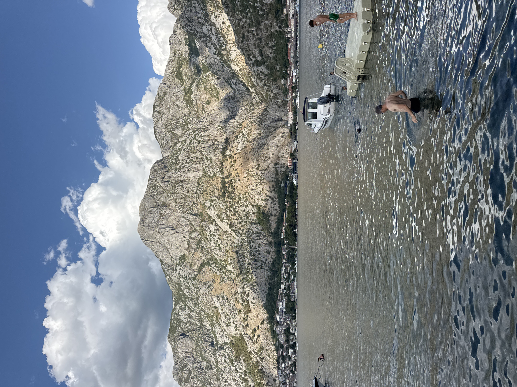 The image depicts a scenic view of the Bay of Kotor, with the town of Škaljari nestled at the base of a steep, rugged mountain. The water in the bay is calm with ripples reflecting the sky and sunlight. Several people are in the water; one man in black swim trunks is wading near the shore, while another, wearing green swim trunks, stands on a floating dock. Two other individuals are partially visible in the water near the dock, and a person is swimming further out. A small white boat with an outboard motor is anchored nearby. The mountain behind the town is covered in sparse vegetation and rocky outcrops, casting shadows on the lower slopes. The sky is a vibrant blue with fluffy white clouds.