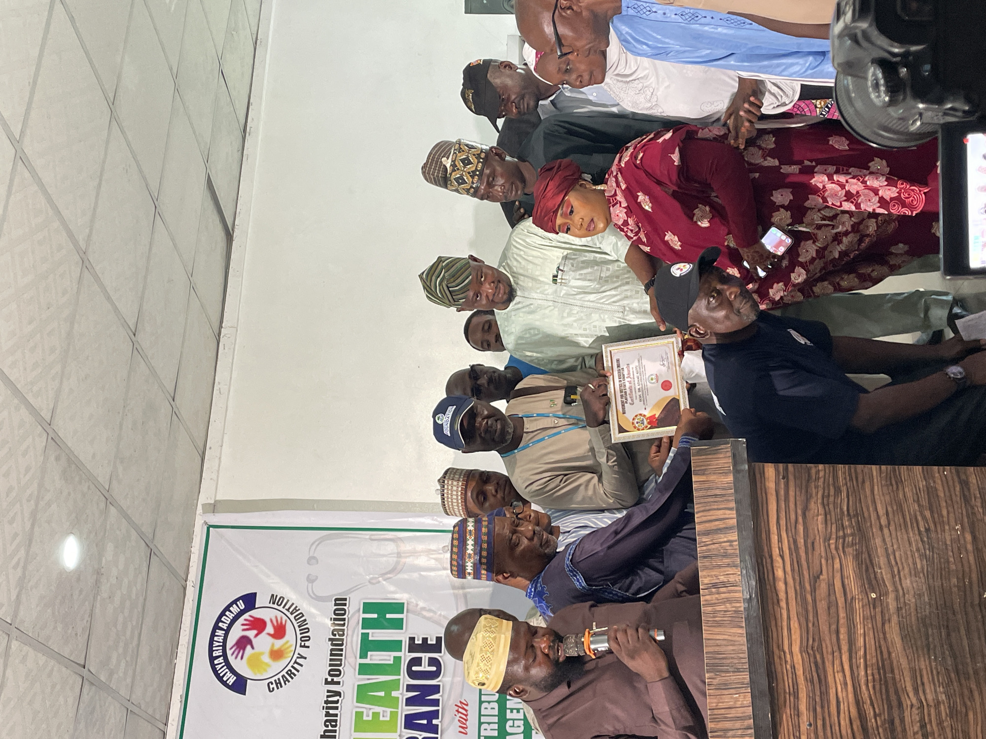 The image captures an indoor gathering in Jos, Nigeria, likely an event organized by the Hajiya Riyan Adamu Charity Foundation. A group of people is positioned behind a banner related to health insurance, and one person is holding a certificate of award with the name "HON. EL ADAM ALIYU" printed on it. Several individuals are dressed in traditional attire. Some are using phones or microphones, and there's a camera recording the event. The environment looks like an indoor event space or office.