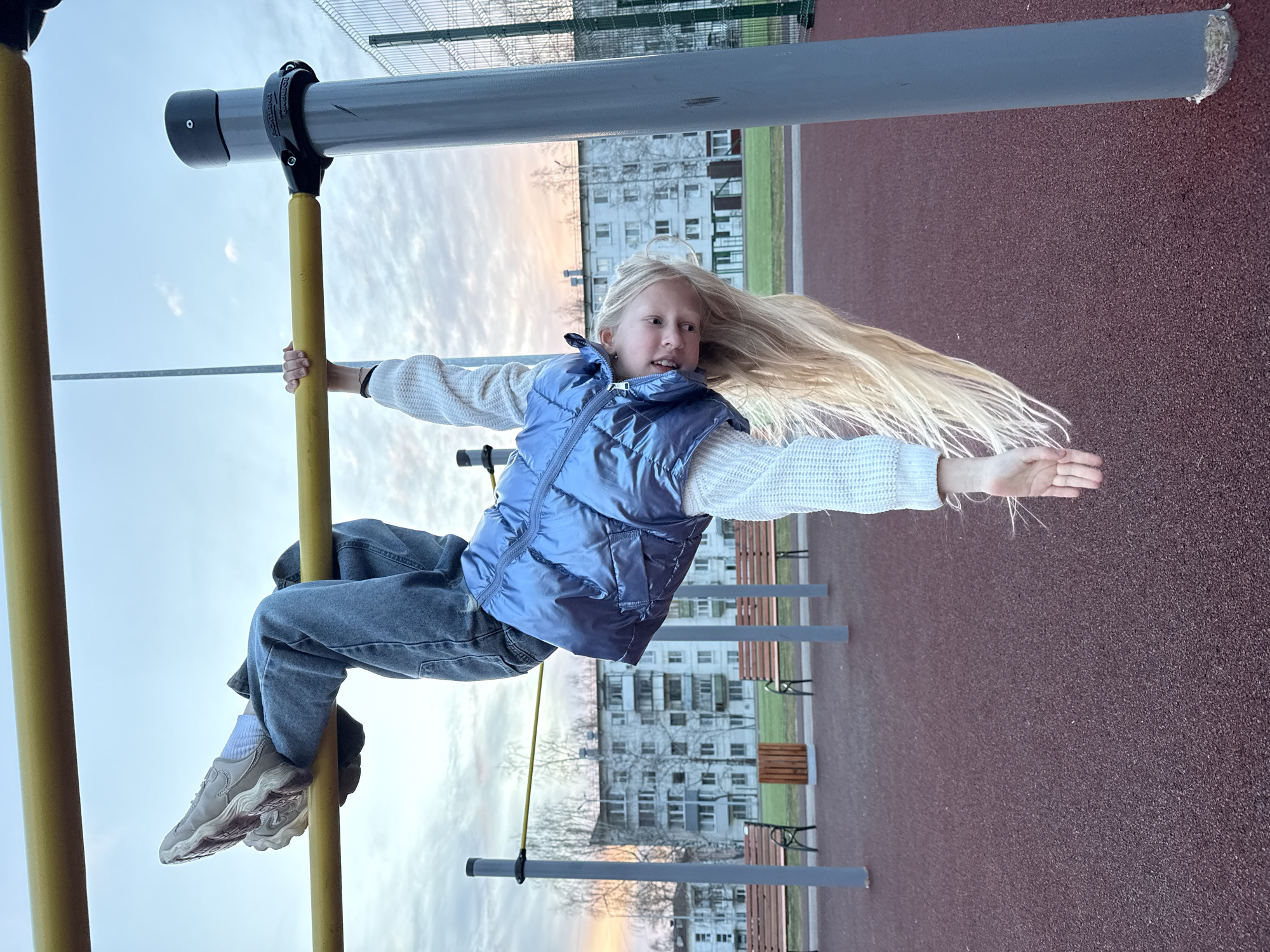 A young girl with long blonde hair is hanging on monkey bars at a playground. She is wearing a light blue vest over a white sweater, blue jeans, white socks, and tan sneakers. She is gripping a yellow vertical bar with her left hand and a yellow horizontal bar with her right hand, her body suspended in mid-air. The playground has a red-brown surface and is partially surrounded by a green metal fence. In the background are apartment buildings, bare trees, and park benches. The sky is visible in the background and appears to be twilight or dawn. The location is in the city of Chaikovsky, Russia.