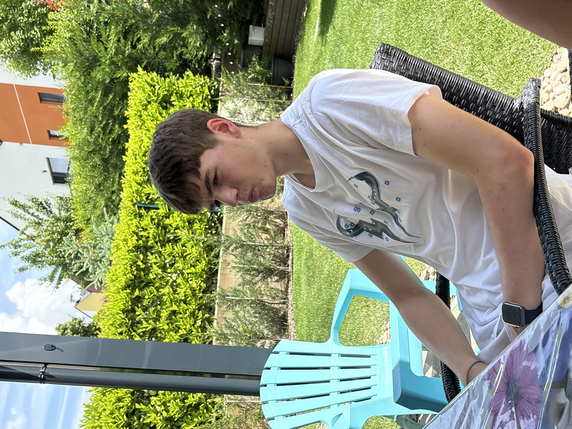 This outdoor image, taken in Lozen, Bulgaria, features a young male seated at a table in a garden on a bright, sunny day. He is positioned in the mid-ground, angled towards the left, and is looking downwards with a focused or contemplative expression. He has short, dark brown hair and is wearing a white short-sleeved t-shirt with a blue and white graphic design on the chest. A dark-colored smartwatch is visible on his left wrist. He is seated in a dark brown wicker chair, and a bright blue plastic chair is partially visible to his left. A table with a clear plastic cover featuring a floral pattern (purple flowers) is seen on the right side of the frame.
The setting is a well-maintained garden with vibrant green grass, numerous lush bushes, and trees under a clear blue sky with a few white clouds. In the background, residential buildings are visible, including one with an orange and white facade. A dark gray metal beam, likely part of an outdoor structure such as a pergola or awning, runs horizontally across the lower left quadrant of the image. The time of day is clearly daytime, indicated by the bright sunshine and clear sky. There is no visible text in the image.
