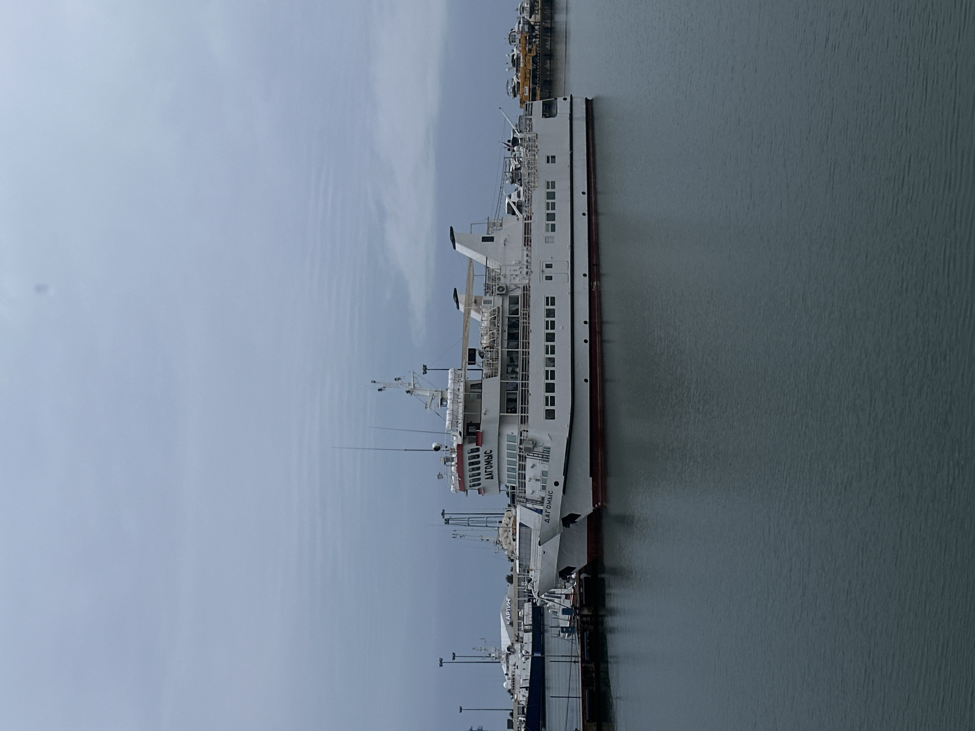 A vertical, medium shot shows the ferry "DAGOMYS" docked in Sochi, Russia, on an overcast day. The white ferry, which has the name “DAGOMYS” in Russian letters on its side, takes up most of the right side of the photo. The ferry is docked next to other boats at a pier. A gray body of water is next to the ship. The sky is pale gray and hazy.
