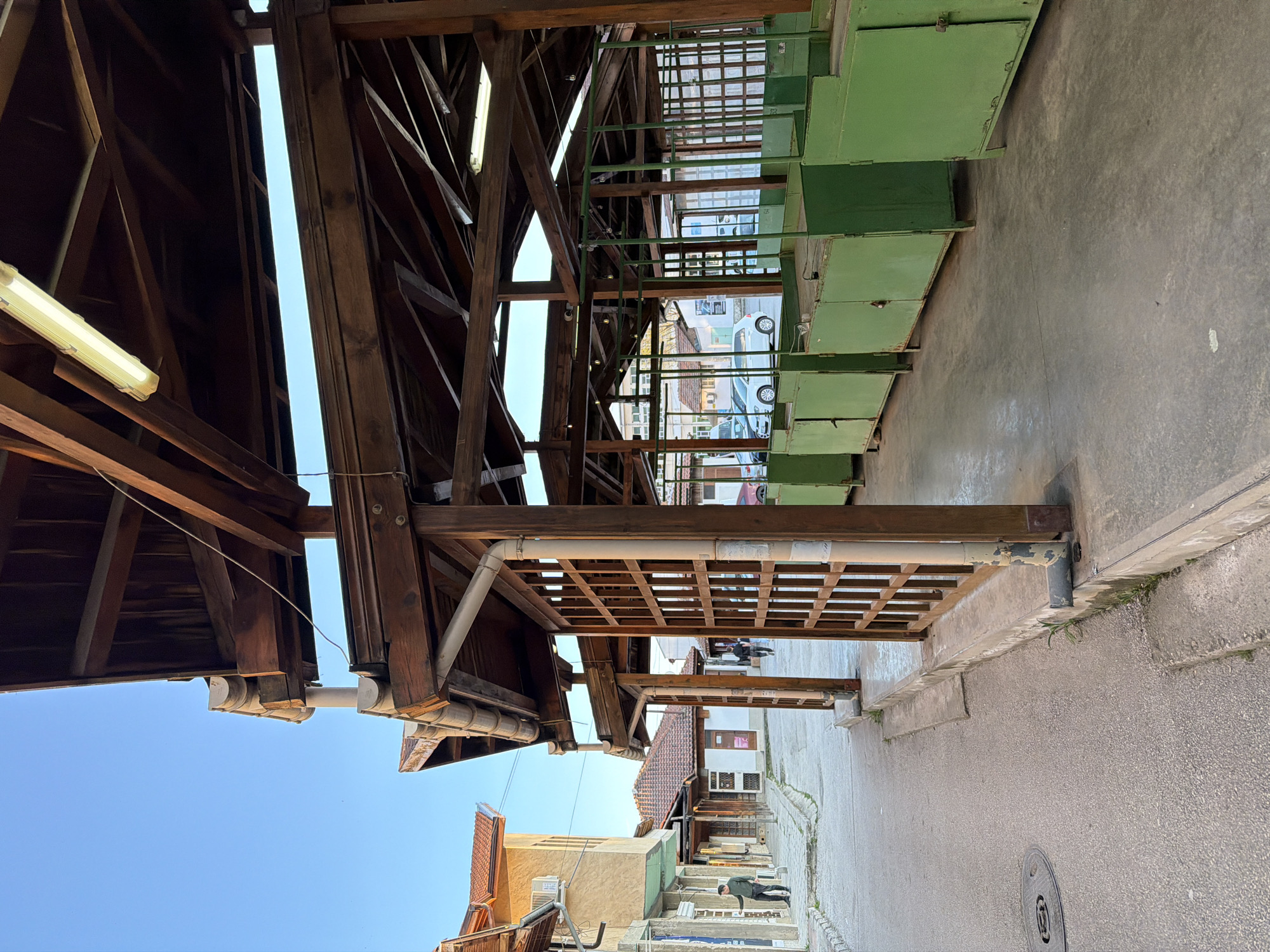 The image depicts an outdoor market area in Panagyurishte, Bulgaria. The foreground features a covered walkway supported by wooden beams and columns. Above the walkway are several rows of green metal storage units or stalls. Beyond the covered area, a street lined with buildings is visible. Two people are walking down the street. The overall scene suggests a traditional market environment within the urban landscape of Panagyurishte.