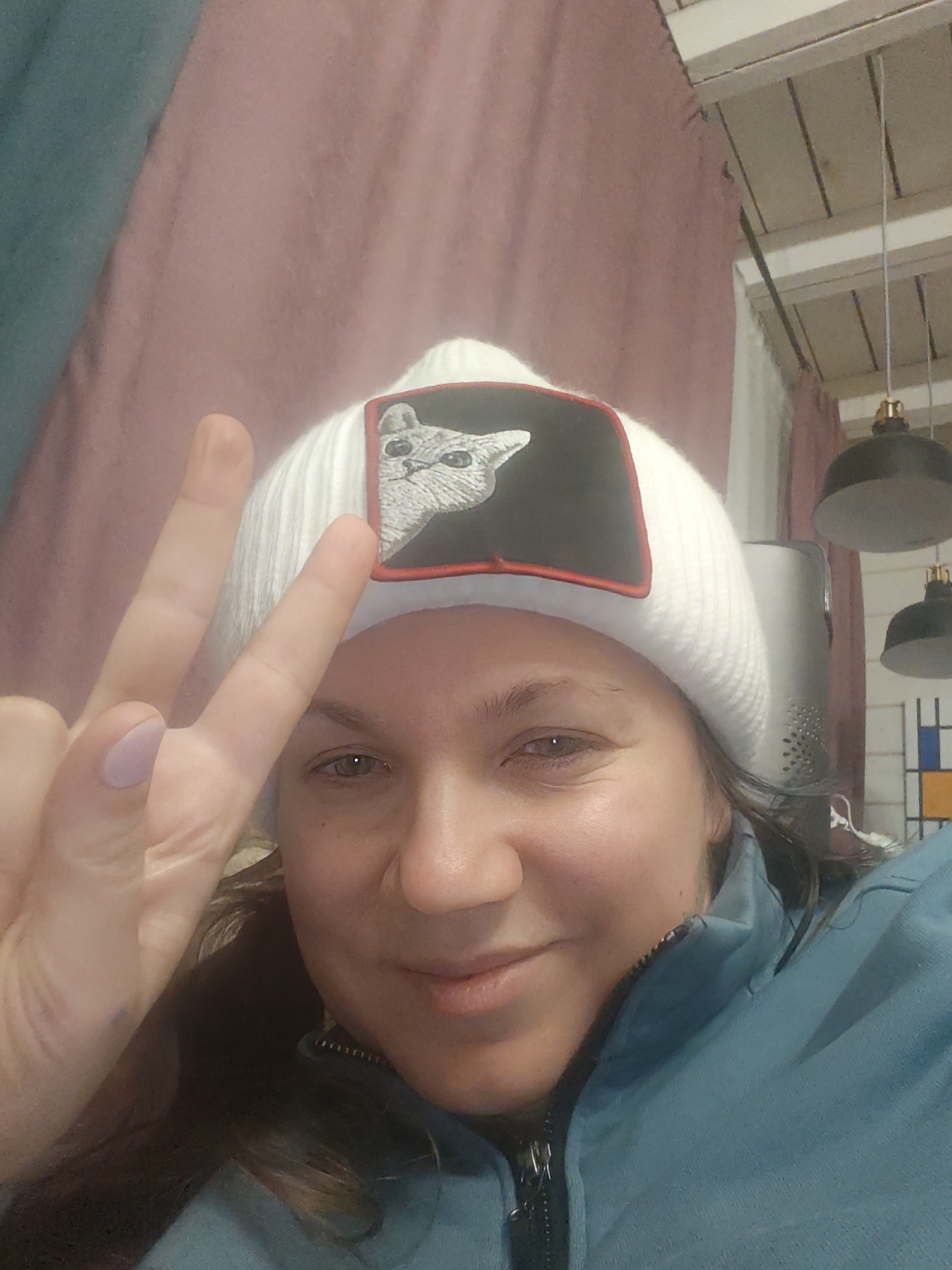 A selfie taken indoors shows a woman wearing a white knit cap with a black patch featuring a white cat. She is making a peace sign with her left hand. She wears a blue jacket, zipped up to the neckline. The background includes a pink curtain, a white ceiling with hanging black pendant lights, and a multicolored abstract art piece in the style of Mondrian. The location is assumed to be in Свистуха (кузяевский С/о), Russia, based on the instructions.
