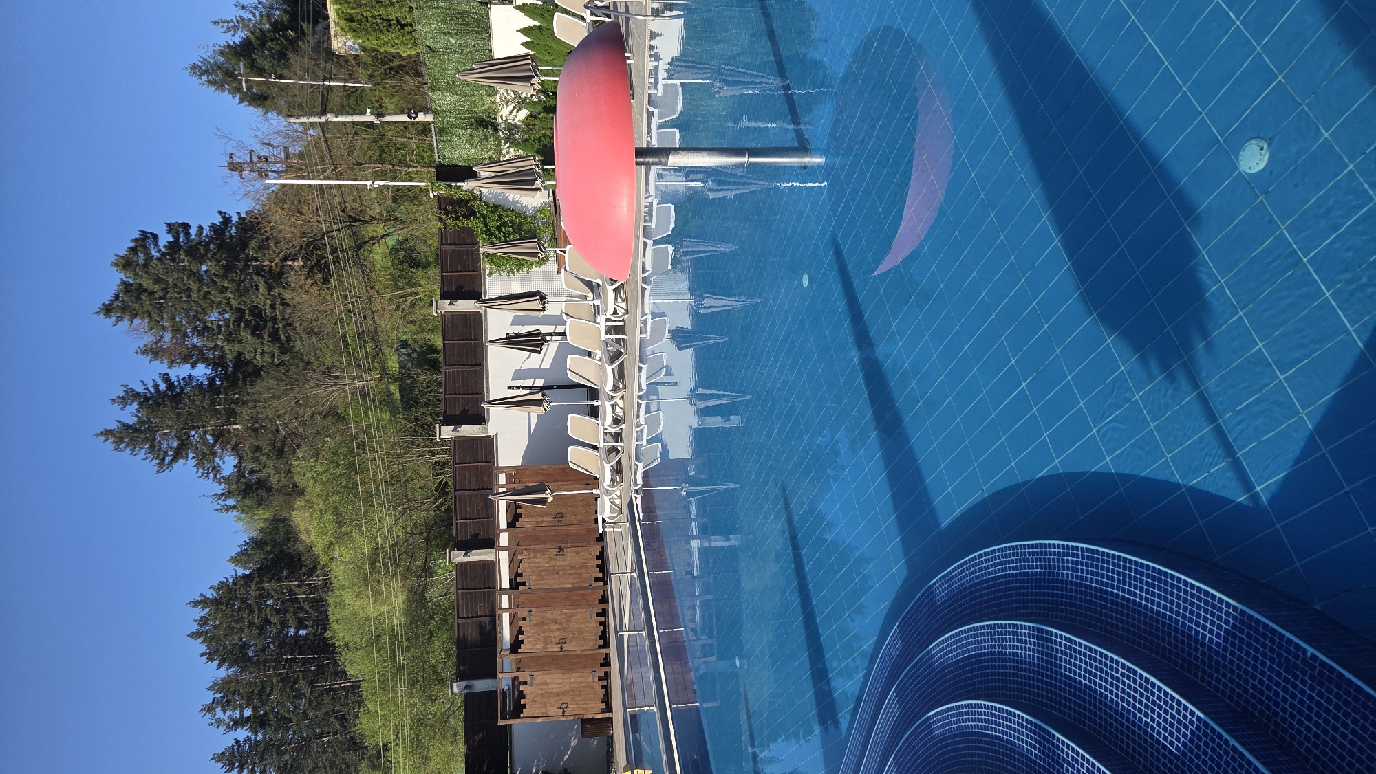 The image depicts a swimming pool area, likely at a resort or hotel, in Unknown, Unknown. The pool's water is a clear blue, and the bottom is lined with blue tiles. A crescent moon design is visible on the pool floor. Lounge chairs are arranged neatly along the poolside, with some shaded by umbrellas. A red object, potentially a kayak or similar watercraft, is positioned near the edge of the pool. A barrier, likely wooden, separates the pool area from a verdant landscape with trees and other vegetation. The sky is a clear blue, suggesting a sunny day. Electrical poles and power lines are visible in the background.