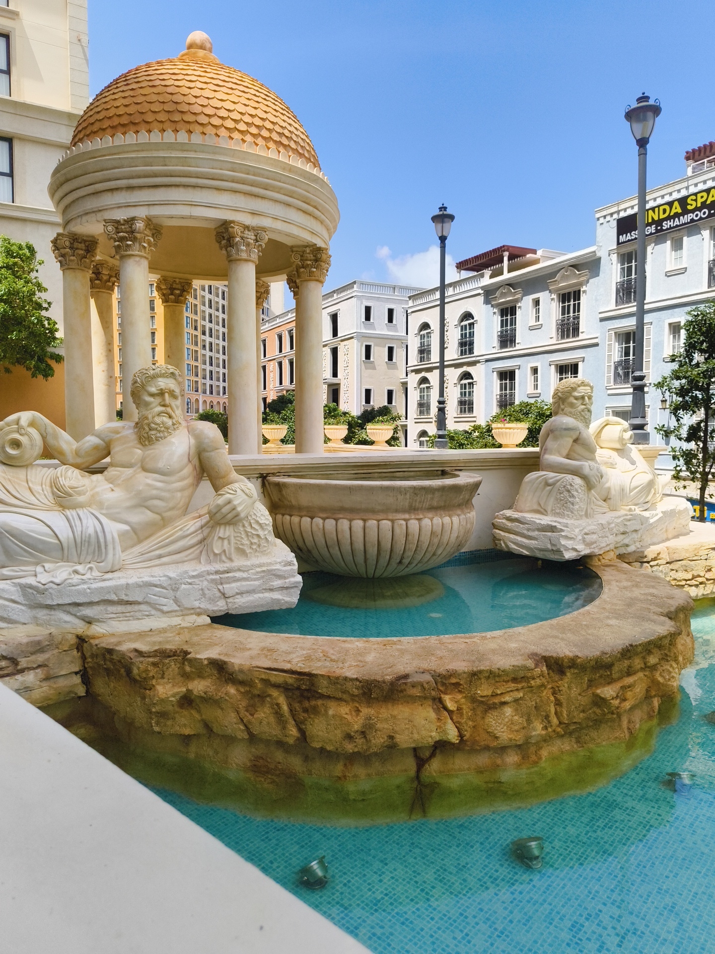 The image depicts a fountain with classical-style sculptures in a city environment. The fountain features two reclining male figures, carved from white stone, positioned on either side of a large, ornate basin. The water in the fountain is a bright turquoise color, with a tiled mosaic bottom. Surrounding the fountain are buildings with a Mediterranean architectural style, characterized by pastel colors, arched windows, and decorative elements. Streetlights are visible in the background, and potted plants line some of the buildings. One building in the background has a banner that reads "VINDA SPA MASSAGE - SHAMPOO". The sky is clear and blue. This scene is located in Phu Quoc, Vietnam, specifically at the Grand World Phu Quoc complex.