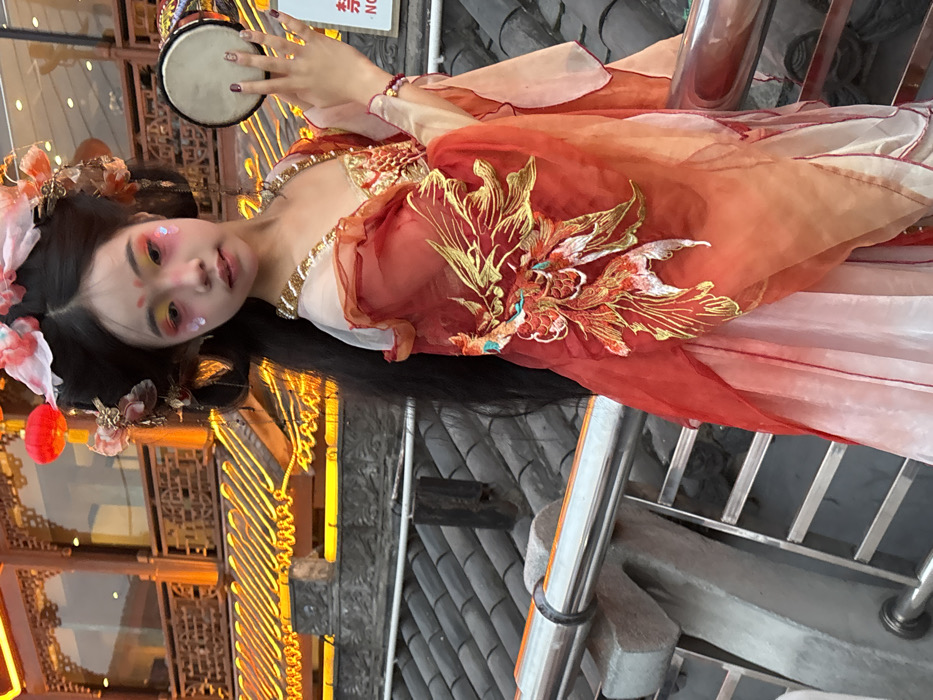 An East Asian individual, appearing young, is depicted from the mid-torso up, positioned slightly to the right of a silver metal handrail with stone supports. The individual faces slightly left of the viewer, looking directly into the camera, and is adorned in an elaborate traditional Chinese costume. The attire consists of a flowing gown in shades of red, orange, and white, featuring gold embroidered phoenix or mythical bird motifs on the left side of the bodice and skirt, and gold trim along the neckline. Long, dark hair is styled upwards with decorative hairpins, pink and white silk flowers, and a red lantern accessory. Dramatic theatrical makeup is applied, including bright orange and yellow eyeshadow, pink facial paint with iridescent accents below the eyes, and small red dots on the forehead. The right hand is raised, holding a small, round, drum-like instrument with a light-colored head and dark rim; fingernails are painted dark. A red beaded bracelet is visible on the left wrist.
The background shows traditional Chinese architecture, likely in Yuzhong District, China. Below the handrail are dark grey, overlapping tiled roofs. Behind the individual, a multi-story traditional building features intricate carved wooden elements and lattice work, illuminated by numerous warm yellow/orange lights outlining the eaves and architectural details. A red Chinese lantern hangs from this structure in the upper left. A red sign with white characters, partially visible with "NO," is in the upper right background. The scene suggests an evening or artificially lit setting.