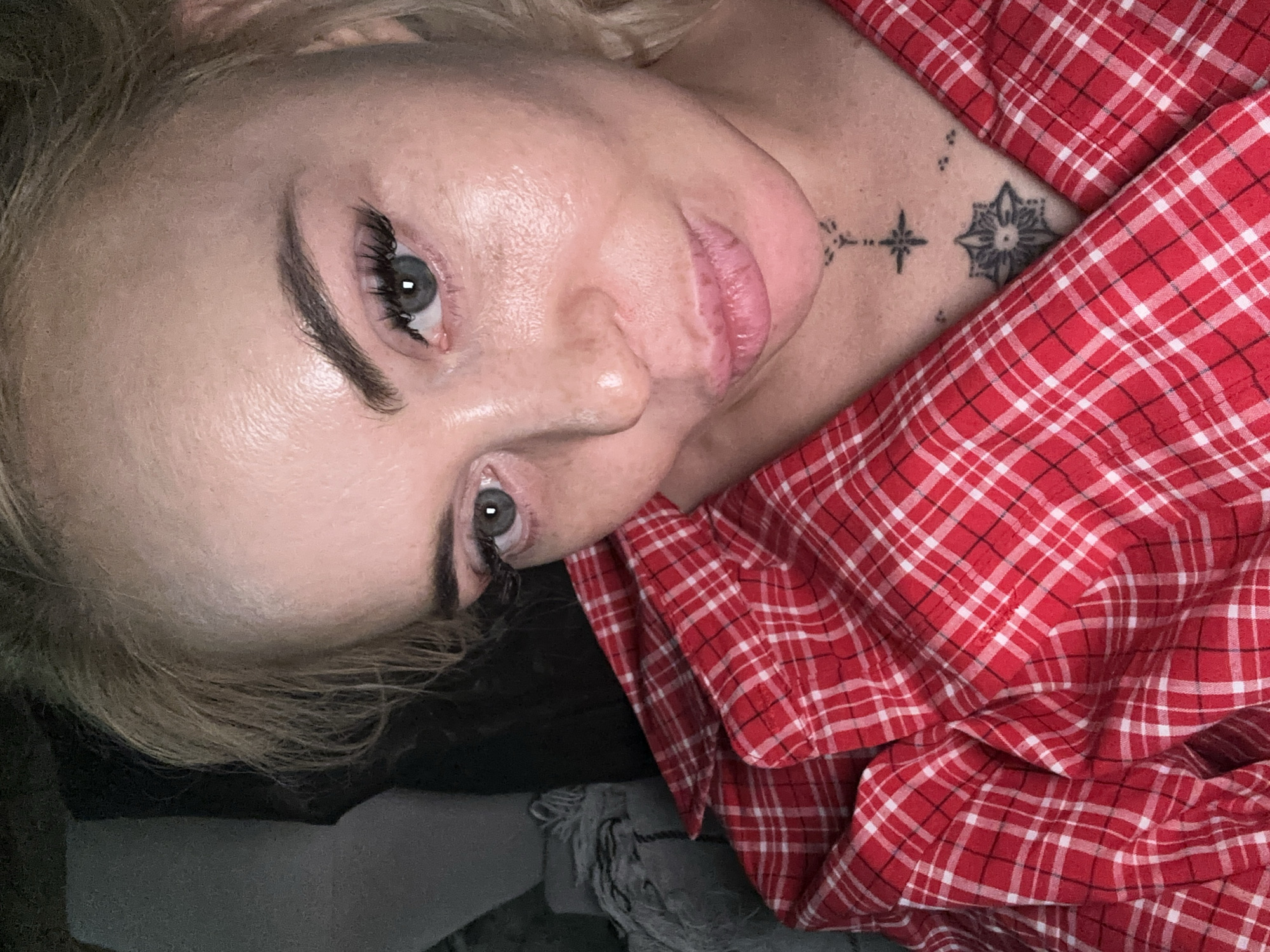 A close-up, high-angle portrait shows an adult individual with blonde hair and light-colored eyes. The individual has a pale complexion with visible skin texture, dark eyebrows, and long eyelashes. The lips are full and appear slightly parted or pouting. Several small black tattoos are visible on the sternum: a stylized cross with dots above it, a four-pointed star, and a circular geometric design below the star. The individual is wearing a red and white plaid collared shirt, unbuttoned at the chest. The background is dark and indistinct, showing a grey, textured fabric object on the left. The specific location in the city of Unknown, Unknown is not discernible from the image.