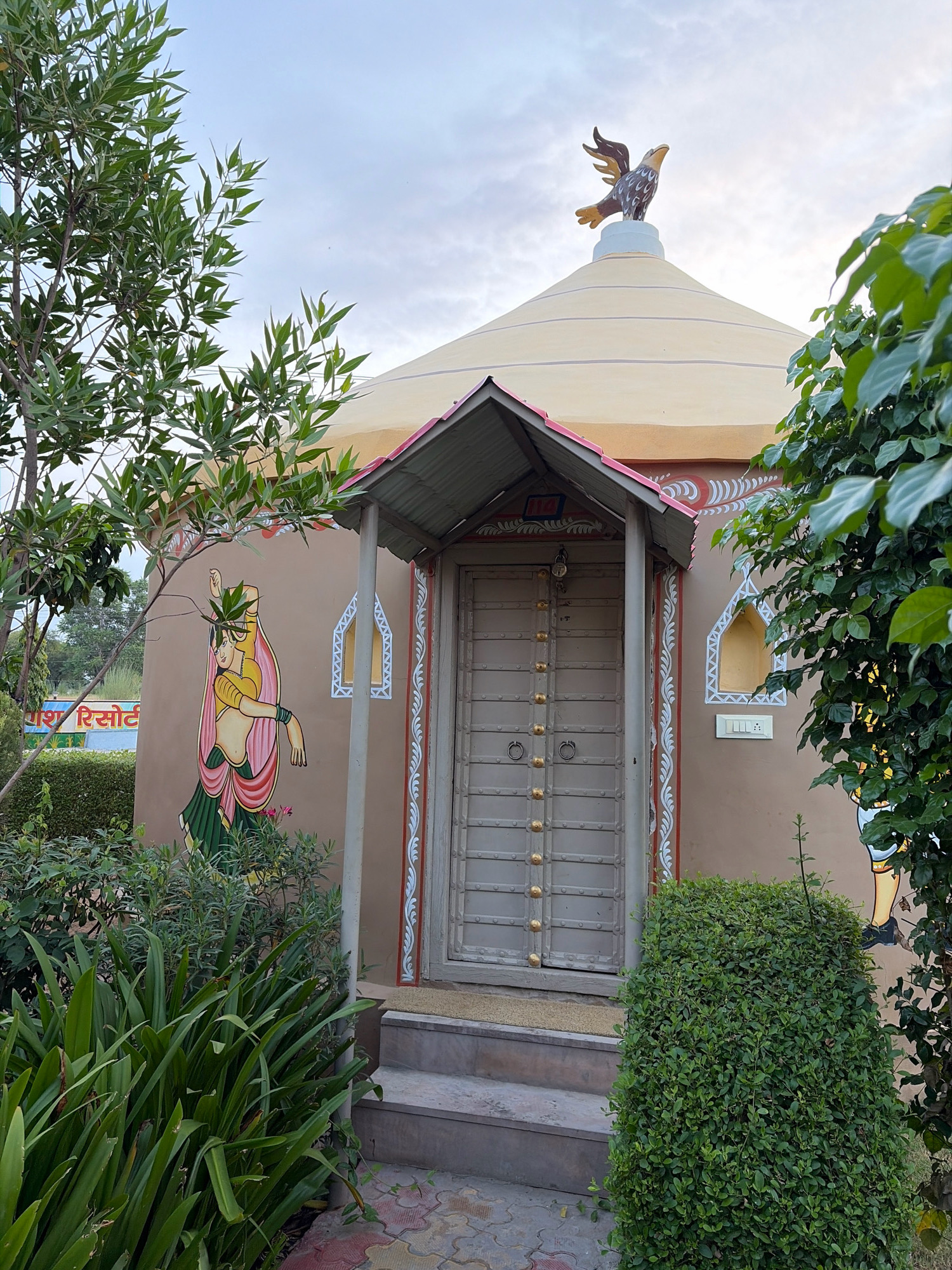 The image shows a small, round building with a conical roof topped with a bird sculpture, likely an eagle, on the exterior in Dockiya, India. The building's walls are painted a light brown and adorned with decorative elements, including a mural of a woman in traditional Indian attire on the left side and white floral patterns around the windows and door. The door itself is a light gray metal door with decorative golden studs and ring handles. A small, covered porch with a corrugated metal roof extends over the entrance, with steps leading up to it. Surrounding the building are various types of greenery, including tall bushes, leafy trees, and a manicured hedge in front of the entrance. In the background, a sign with Hindi text, possibly indicating a resort, is visible. The sky is partly cloudy, suggesting it is either early morning or late afternoon.