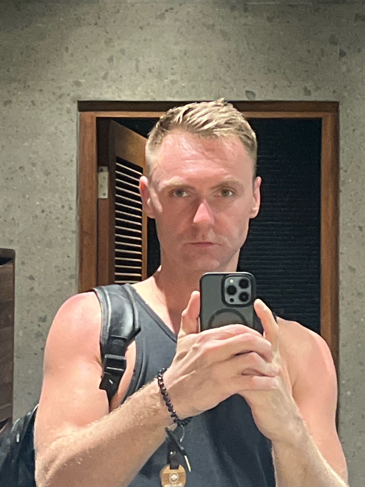 A man with short, blonde hair and a muscular build takes a selfie in a bathroom. He is wearing a black tank top and a black backpack strap is visible over his shoulder. He is holding a smartphone in his right hand, and the phone's camera is pointed at himself. The background shows a textured grey wall and a wooden door with louvers. The lighting is bright and appears to be artificial. The man's gaze is directed at the camera.