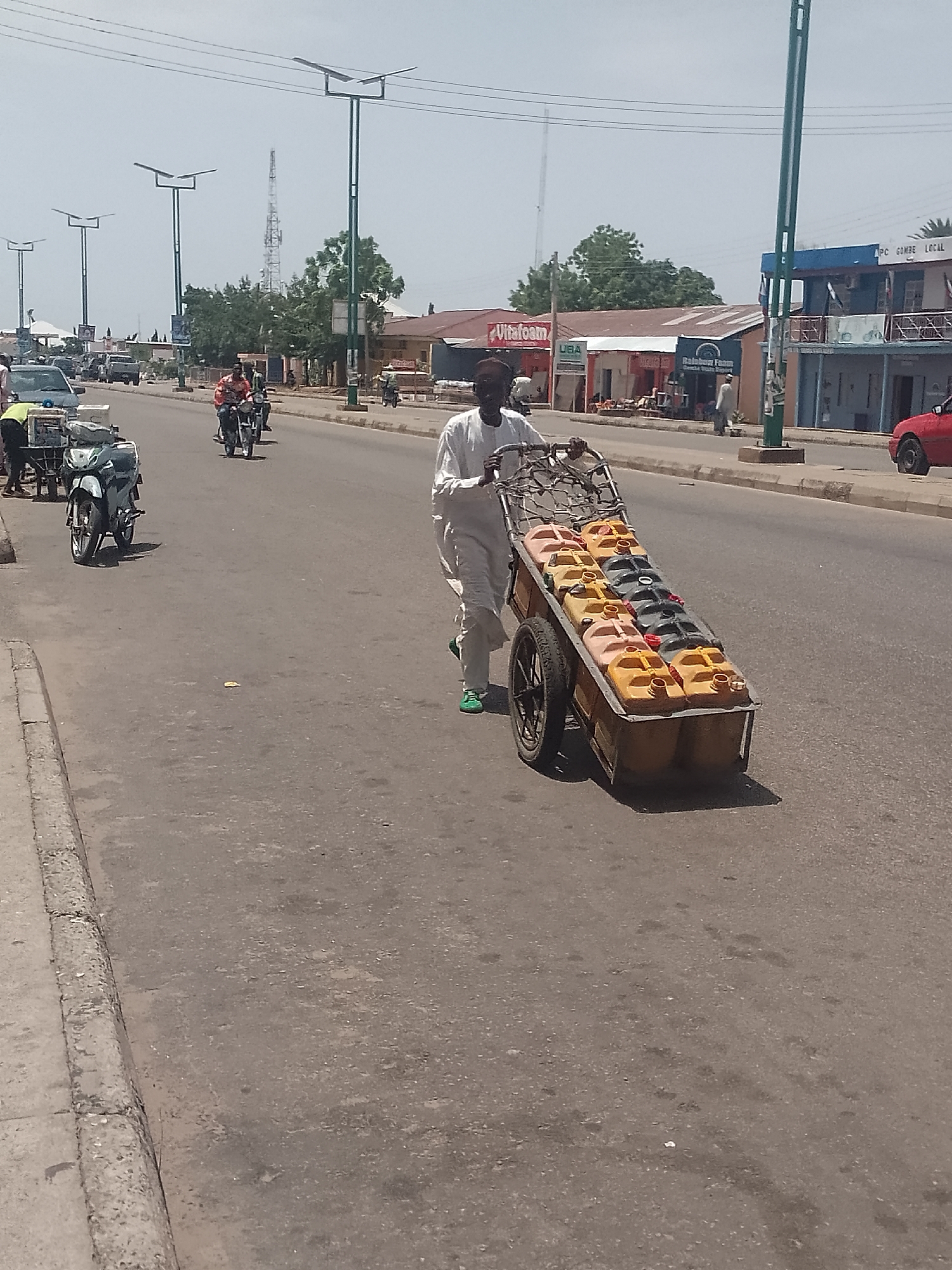 A man in a white outfit and green shoes is pulling a two-wheeled cart filled with rows of yellow and black plastic containers along a paved road. Buildings are visible in the background, including one with a "Vitafoam" sign, and another with "APC Gombe LOCAL" displayed. The scene is set on a sunny day in what appears to be an urban area, indicated by the presence of street lights, vehicles, and other pedestrians. A motorcycle and other vehicles are on the road.
