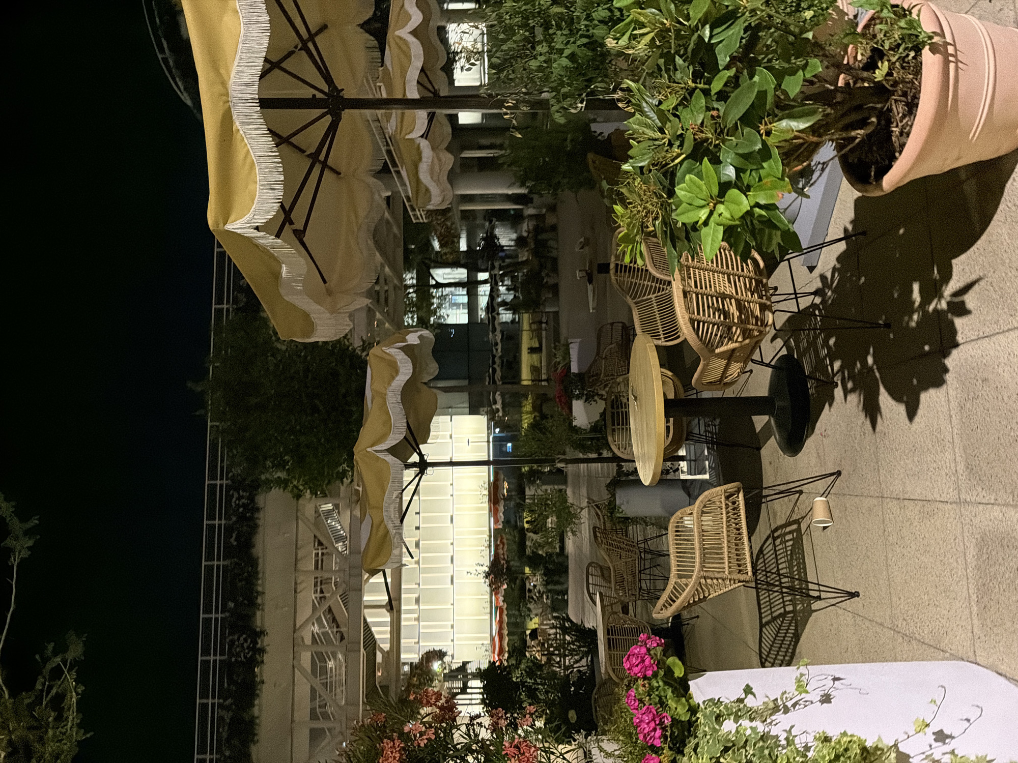 The image depicts an outdoor seating area of a cafe or restaurant at night, likely in Moscow, Russia, given the context of previous similar images. The scene is illuminated by artificial lights, casting long shadows. In the foreground, there are several rattan chairs and round tables. One table is a light wooden top with a dark base, and another is a dark circular table with a light top. A terracotta pot containing a large green plant is visible on the right. In the background, more tables and chairs are set up under large, scalloped yellow umbrellas with white fringe. The building in the background has a modern facade with illuminated horizontal bands. There are no people clearly visible in the immediate foreground. A disposable cup is lying on its side on the ground near a chair. The overall atmosphere suggests a tranquil, nocturnal outdoor dining setting.