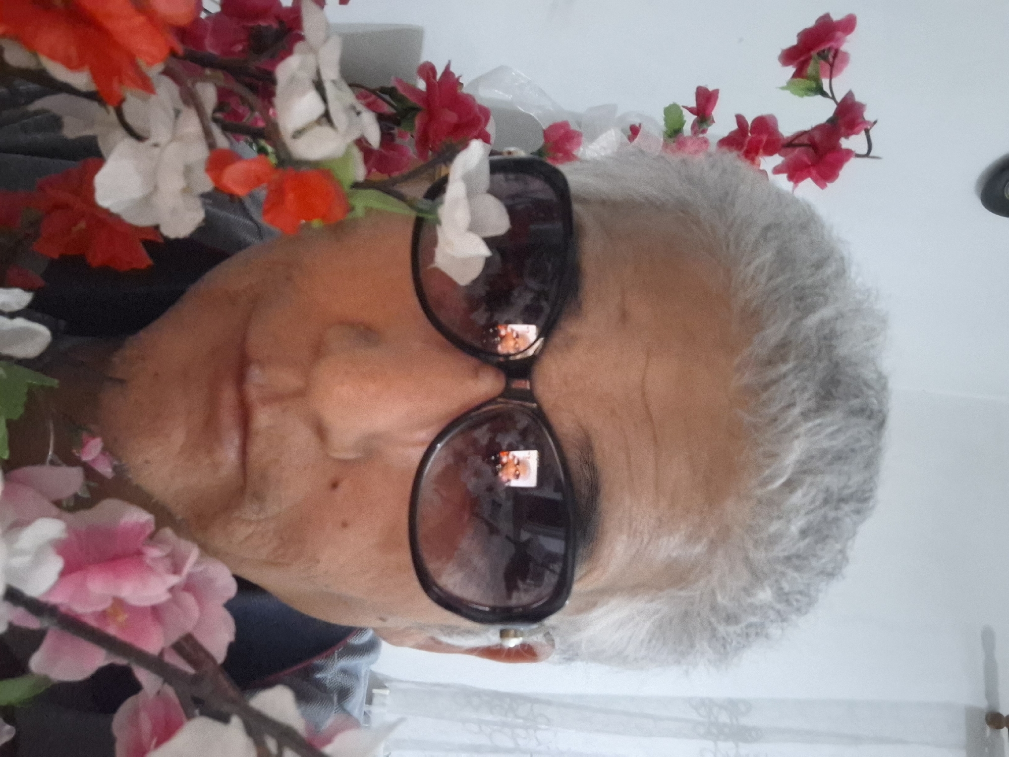 The image is a close-up, selfie-style photograph of an older person with gray hair, wearing large, dark-rimmed sunglasses. Artificial flowers, in shades of pink, white, and red, are arranged in the foreground, partially obscuring the subject's face and chest. The subject is looking directly at the camera. Reflections of the subject, and potentially another small figure, are visible in the lenses of the sunglasses. The background appears to be an indoor setting with a white wall and sheer white curtains. The provided information does not specify a location in Almaty, Kazakhstan.