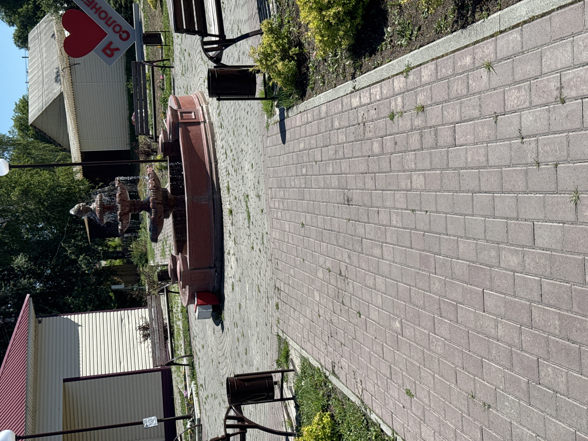 The image shows an outdoor scene with a paved walkway leading towards a fountain. In the background, there are buildings, trees, and park benches. A prominent sign with a red heart and the inscription "Я СОЛОНЕ" is visible. The environment appears to be a park or public square in the city of Soloneshnoye, Russia. There are no people visible in the image. The lighting suggests it is daytime, with sunlight casting shadows. The overall scene is tranquil and well-maintained.