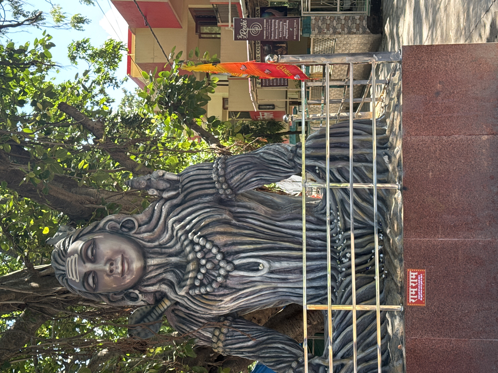 A large, detailed statue of the Hindu deity Shiva, made to resemble bark and wood, is situated outdoors. The statue faces forward, with its eyes closed and a serene expression. It is adorned with a necklace of beads and has a trident symbol on its forehead. To the right of Shiva's head, a smaller statue of a child is visible. The statue is partially shaded by the dense foliage of a large tree. A metal railing separates the statue from the foreground. In the background, there is a multi-story building with a red and yellow facade, and a vibrant orange and yellow flag is displayed on a pole. A sign for "Reiki" and "BRAHM YOGA" is also visible on the building. At the base of the statue, on a reddish-brown textured wall, a small sign with Hindi text that reads "राम राम" is affixed. The scene appears to be a public space, possibly a temple or shrine, in Rishikesh, India, given the cultural and religious iconography.