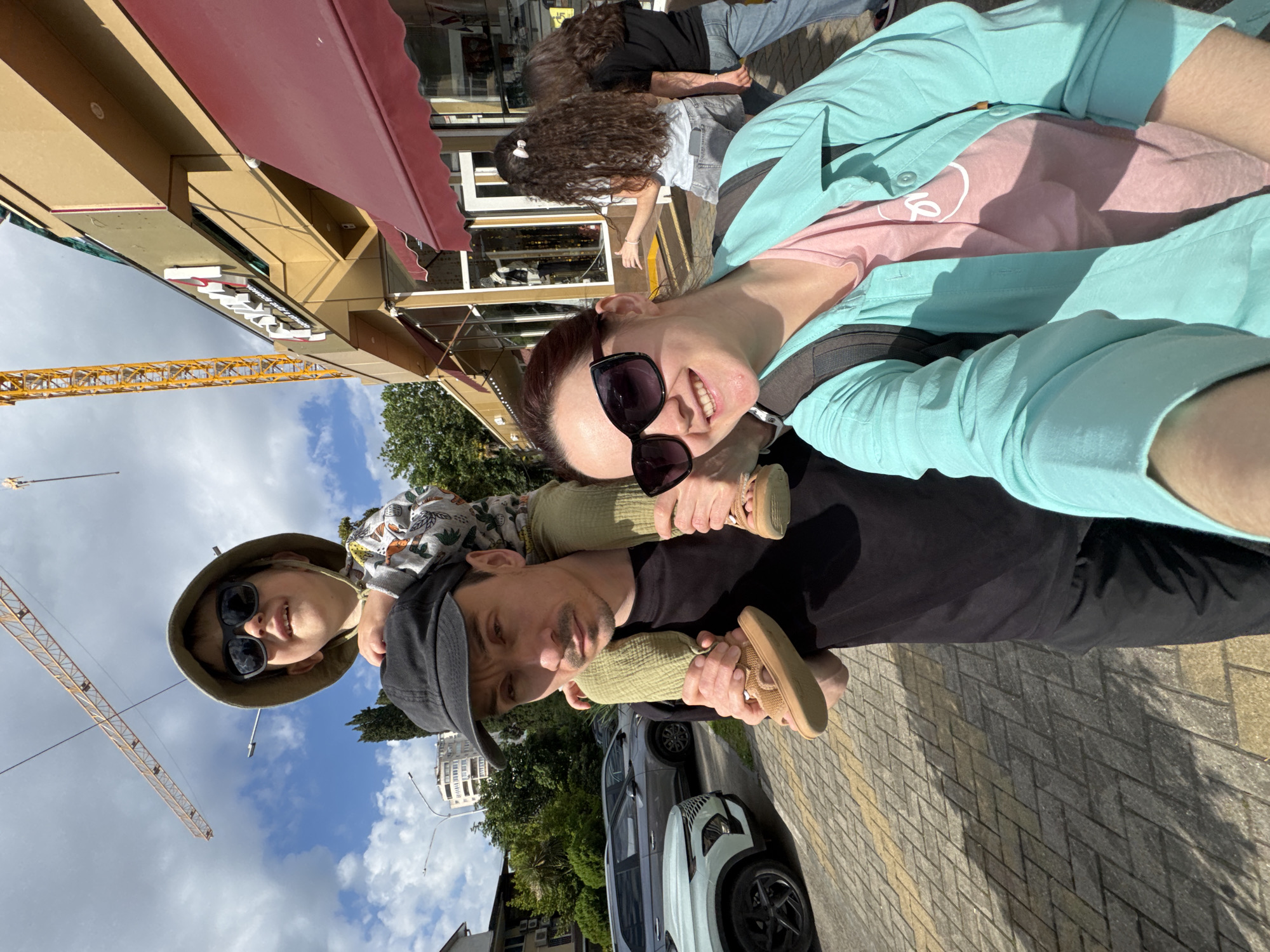 The image captures a family selfie in Sochi, Russia. A father carries his child on his shoulders, while the mother holds the camera. The child wears a hat and sunglasses, while the parents also wear sunglasses. A crane is visible in the background, suggesting construction is ongoing in the area. Pedestrians walk behind the family on the sidewalk. Cars are parked nearby. The photo was taken during the day, evidenced by the bright sunlight and shadows. The location appears to be a city street with buildings and trees.