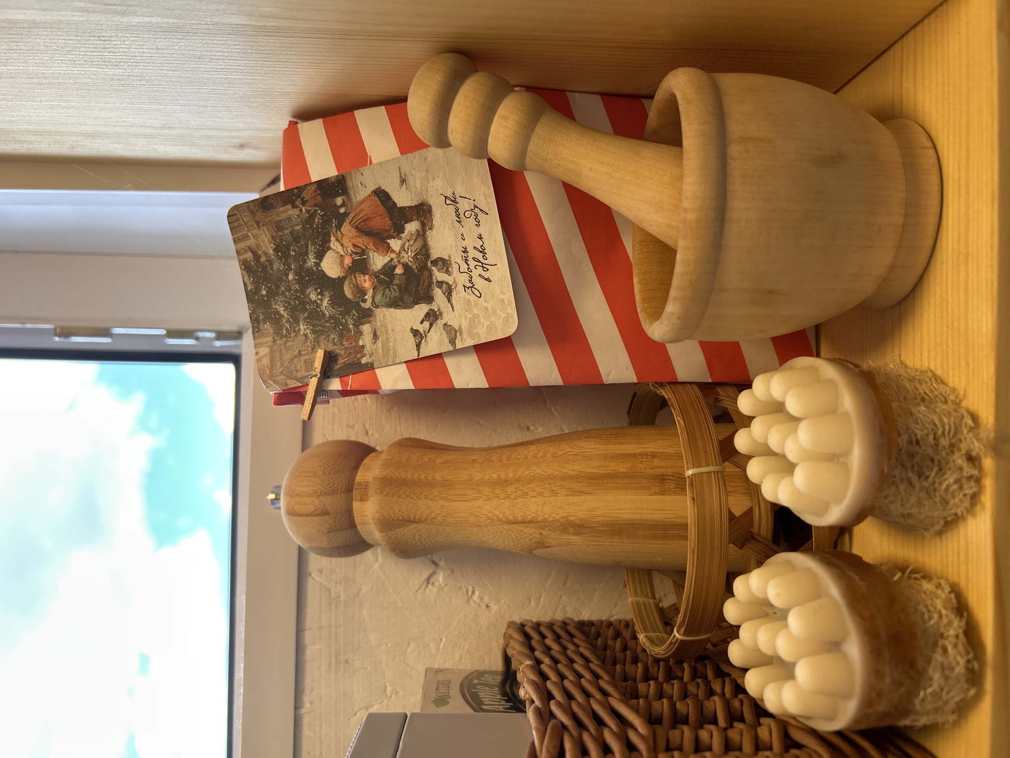 This media file depicts a collection of items placed on wooden shelves. The main subjects are a wooden mortar and pestle, a striped red and white gift wrap, and a postcard. In the foreground, a wooden massage roller sits within a woven basket, flanked by two round loofah sponges. The scene is indoors, with a window showing a bright blue sky with white clouds, suggesting daytime.
The postcard features an illustration of two children in winter attire, seemingly interacting with pigeons in a snowy setting. Some Cyrillic text is visible on the postcard, which reads "Заботы с Любовью в Новом году!" (Care with Love in the New Year!). The gift wrap is bright red and white, with diagonal stripes.
Below the massage roller, a woven basket is visible, and a small package with text that includes "Apple Tea" and "NATURE" is partially seen. The overall impression is one of a curated collection of decorative and possibly functional items, perhaps for a spa or a gift display. The lighting suggests an indoor environment with natural light from a window. The scene appears static, with no active event taking place.