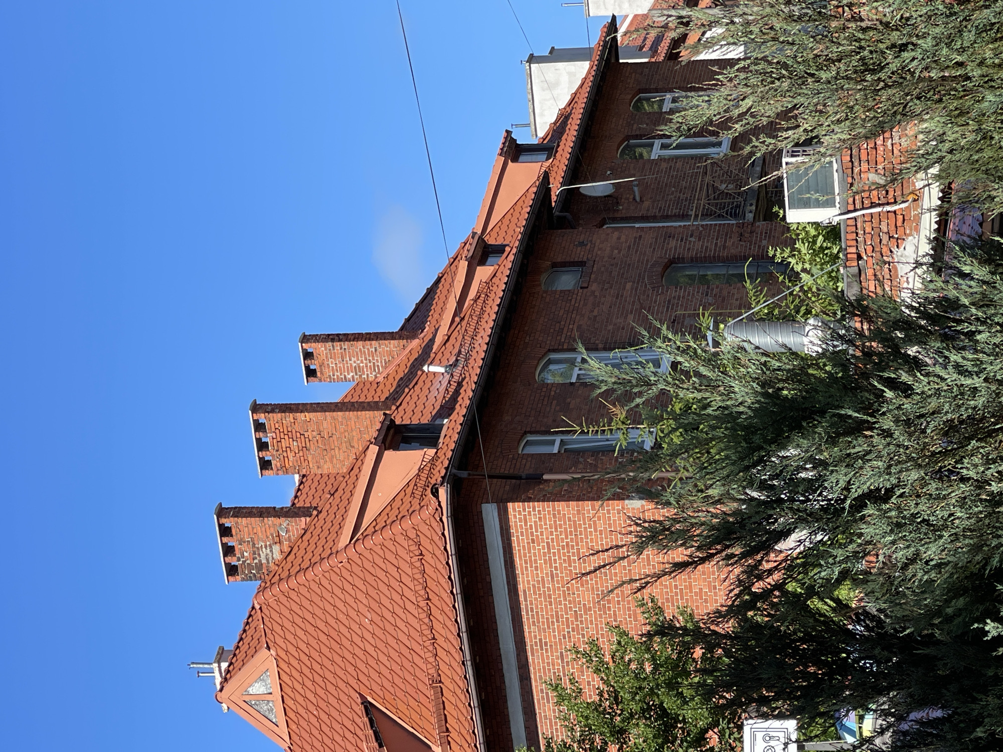 This is a medium-shot, eye-level, outdoor, daytime, full-color photograph of a brick building with a red tile roof. The building is located in the city of Zelenogradsk, Russia, and is partially obscured by dense green coniferous trees in the foreground. The sky above is a clear, bright blue with a few wispy white clouds.
The building itself appears to be an older, multi-story structure. The facade is made of red brick, and several windows with white frames are visible. The roof is steep and covered in red, overlapping tiles, with multiple brick chimneys emerging from it. One chimney on the left has a visible metal flue pipe attached. There are dormer windows on the roof as well. A small, triangular window with a light-colored frame is set into the front gable of the roof on the left side of the image. On the right side of the building, there is a small balcony with a metal railing. An air conditioning unit is visible on a lower roof section on the right. Wires are strung across the sky above the building. In the distance, the tops of other buildings, some with white exteriors, are visible.