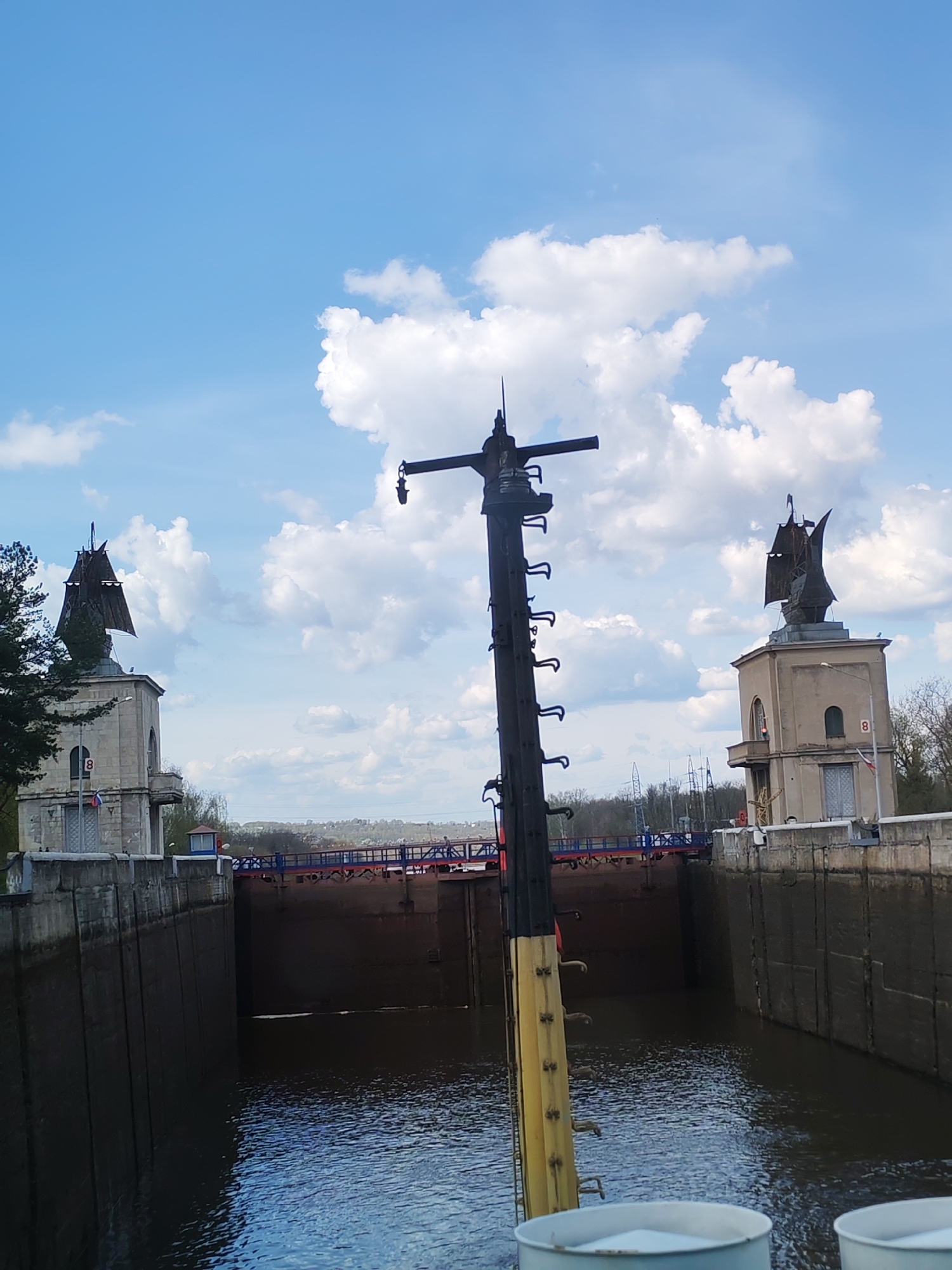 The image shows the lock of the Moscow Canal in Yakhroma, Russia. The lock chamber is framed by concrete walls. At each end of the chamber, there is a control tower topped with a sculpture of a sailing ship. A blue and red bridge spans across the locks in the background. A tall, black and yellow pole, likely part of a vessel, is prominently positioned in the foreground. The sky above is blue with scattered white clouds. The water in the lock is dark and reflective.
