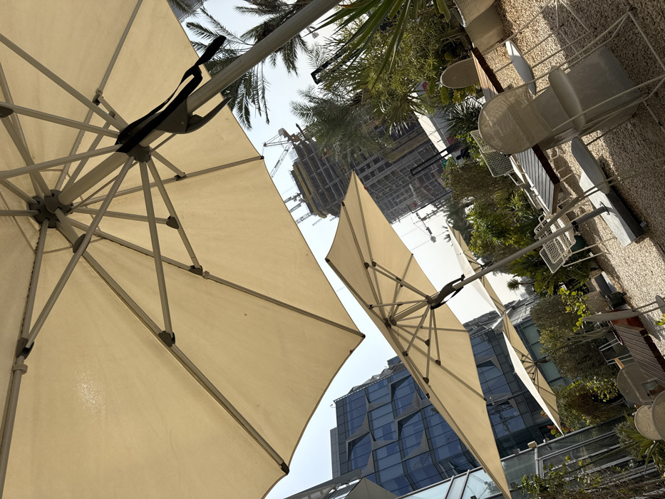 This image depicts an outdoor cafe or lounge area located within Dubai Design District (d3), Dubai, United Arab Emirates. The scene is observed from a low angle, looking upwards and across the space.
In the foreground, several large, open patio umbrellas with beige fabric canopies are prominently featured, seen from underneath, revealing their white metal frames and support structures. The closest umbrella dominates the upper portion of the frame. Below these umbrellas, the ground is covered with light-colored gravel or small pebbles.
Multiple outdoor seating arrangements are visible, consisting of modern tables with light-colored wooden tops and white or light grey metal chairs, some featuring a mesh or woven design. These tables and chairs are unoccupied. Various green plants, shrubs, and small palm trees are interspersed throughout the area, with some plants contained within white cylindrical planters.
The background reveals urban development under a clear, bright sky. To the left, a modern architectural building with a distinctive blue, reflective glass facade showcasing geometric patterns is partially visible. To the right, a tall building is under active construction, identifiable by its exposed concrete structure, scaffolding, and multiple yellow tower cranes at its summit. No individuals or specific interactions are present in the depicted scene.