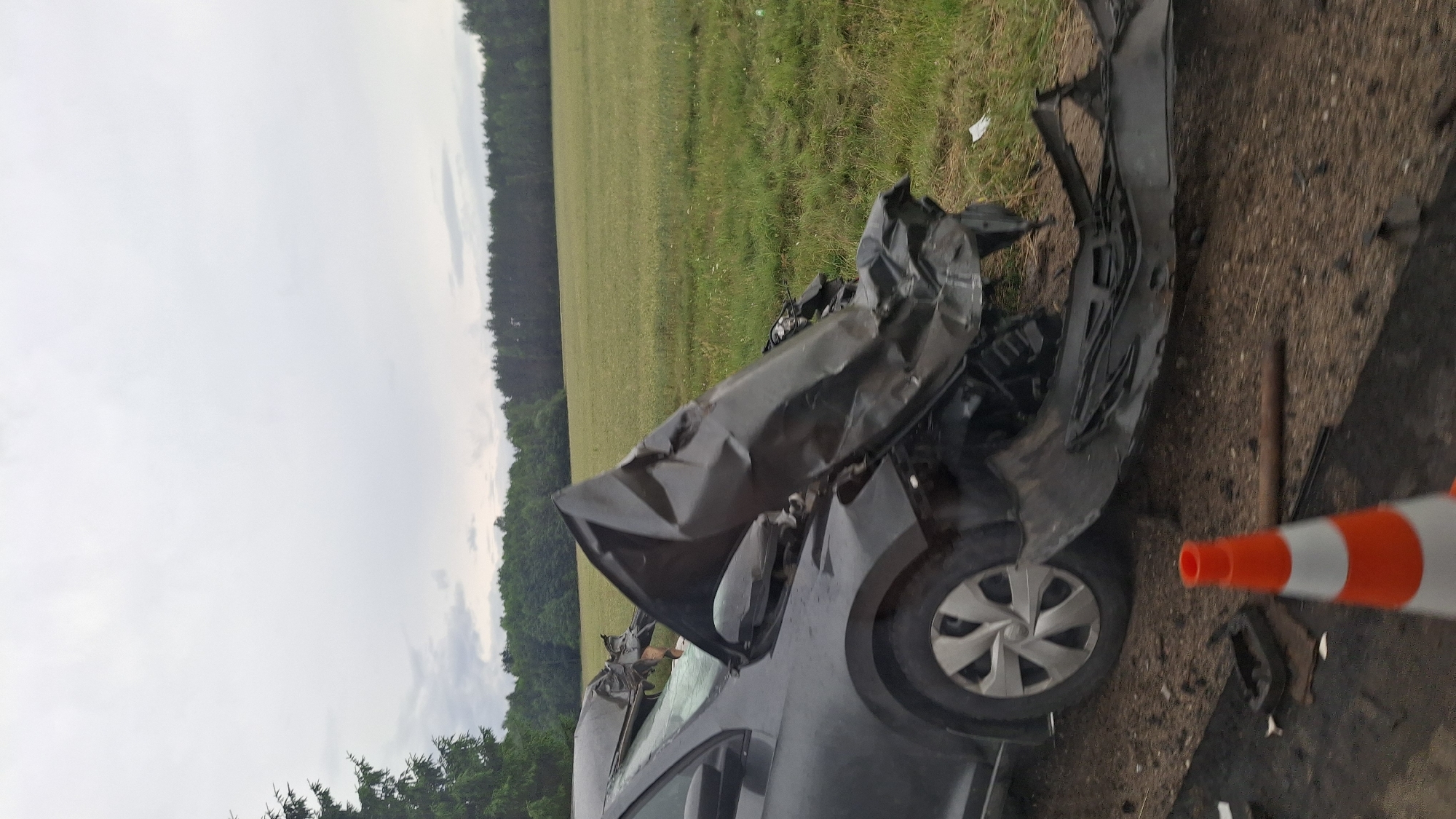 A dark-colored, severely damaged car is situated on the side of a rural road. The front of the vehicle is crumpled and deformed, with parts of the hood and bumper detached and bent outwards. The car is angled, with its front left wheel clearly visible and its rim intact. A small orange and white traffic cone is positioned in the foreground, near the car's front tire. The background consists of a grassy field bordered by a dense line of trees under an overcast sky. The setting appears to be outside a populated area, consistent with a location in the countryside near Ilyanskiy selskiy Sovet, Belarus. There are no people visible in the image.