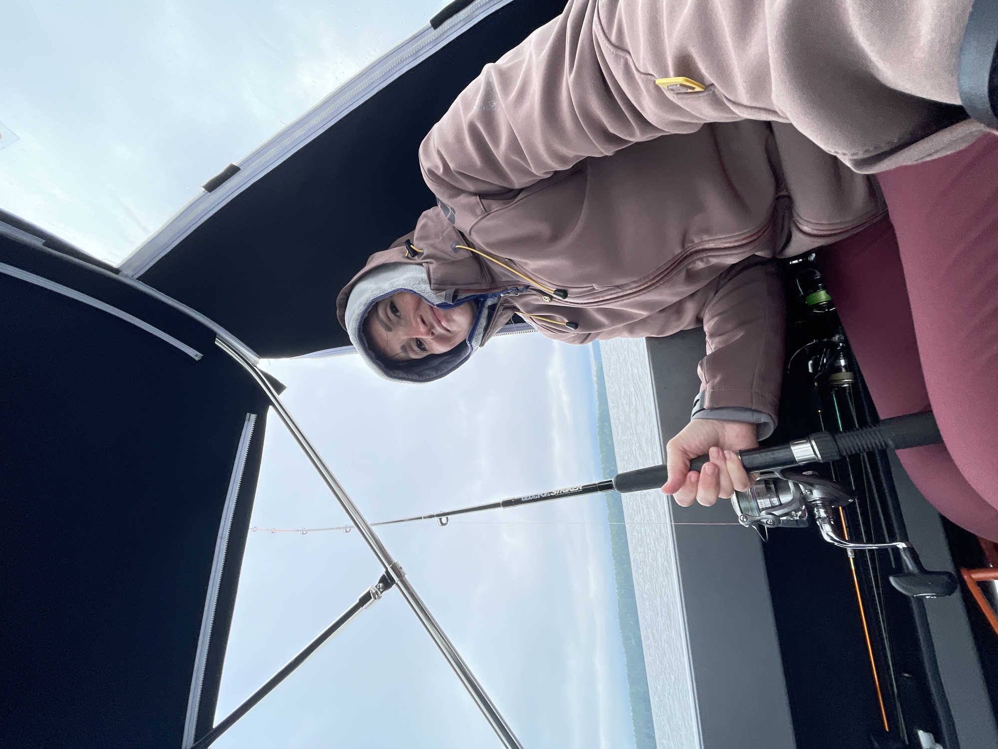 A woman is fishing from a boat on a cloudy day in Yablonovka, Russia. She is wearing a brown hooded jacket and burgundy leggings. She is holding a fishing rod with a silver reel. The boat has a dark blue canopy. The water is calm and there is a wooded shoreline in the distance.