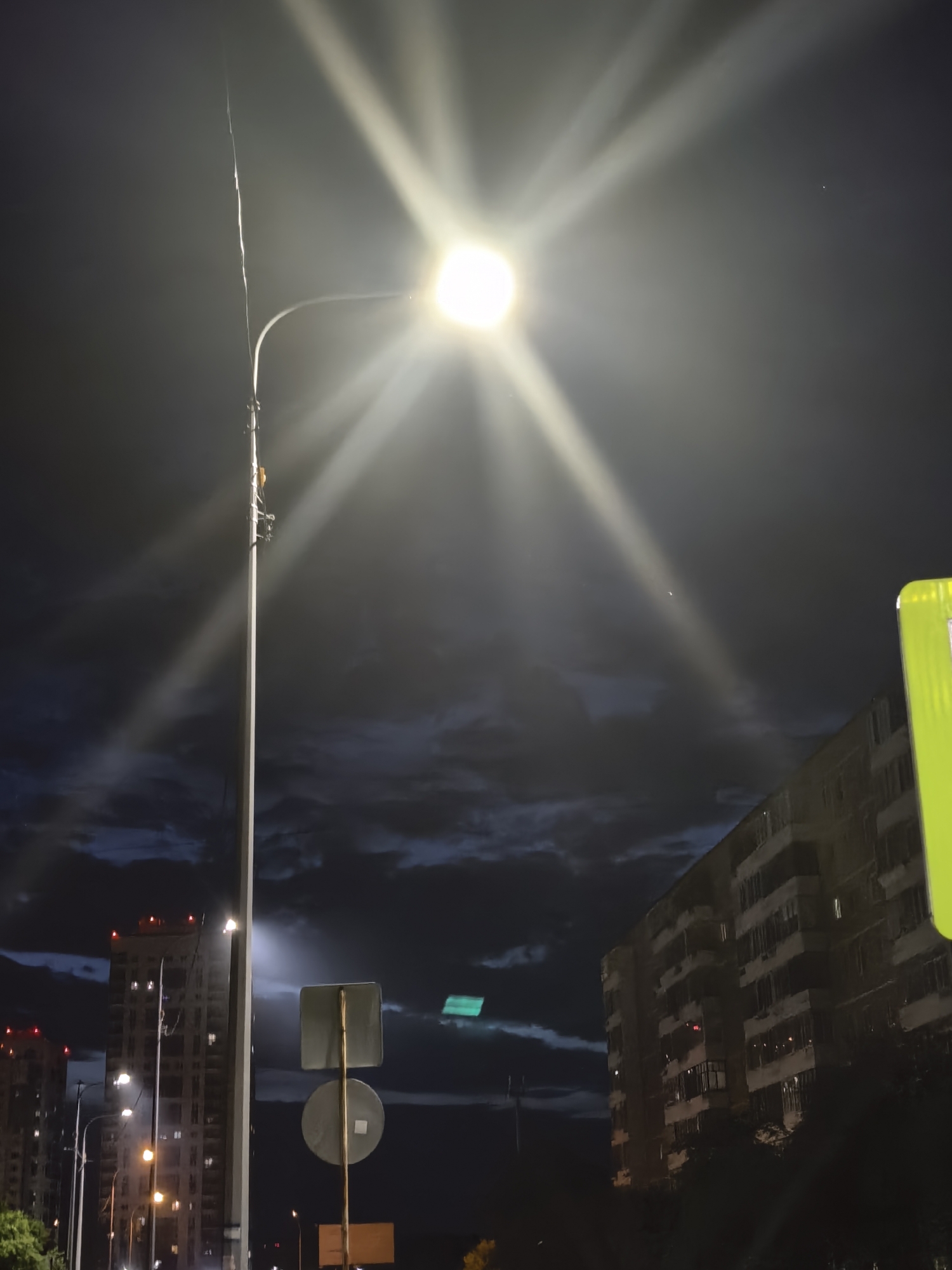 An eye-level, wide-angle shot captures a streetlamp illuminating a dark, cloudy night sky over a city. The streetlamp, positioned in the left half of the frame, emits a bright, starburst-like light, with beams extending outwards and creating lens flares. Below the streetlamp, a traffic sign with two circular signs is visible. To the right of the streetlamp, a building with multiple balconies and windows is partially lit by the lamp. In the background, other buildings with illuminated windows are visible, along with streetlights. The clouds in the sky are dark and appear textured, suggesting movement. The overall scene is illuminated by the streetlamp and ambient city lights, creating a moody and somewhat dramatic atmosphere.