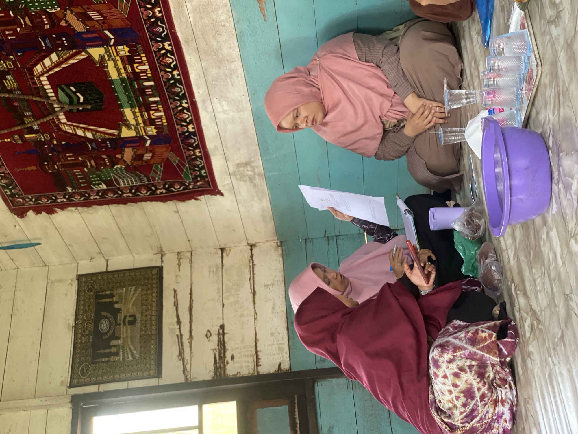 Three women wearing hijabs are sitting on the floor of a room with wooden walls. The room is sparsely furnished and appears to be a simple dwelling. One woman on the right is wearing a pink hijab and brown clothing, and she is looking to the left. In the center, another woman in a pink hijab is looking down at a piece of paper held by a third woman on the left. The woman on the left is wearing a maroon hijab and a patterned skirt, and she is holding a mobile phone in her left hand. In front of the women are various objects, including plastic bags, plastic cups, a purple bowl filled with water, and a tray with several glasses. Two religious tapestries hang on the walls; one depicts the Kaaba in Mecca, and the other is a prayer rug with an image of the Kaaba and surrounding buildings. The floor has a marble-like pattern. The scene suggests a casual meeting or gathering, possibly for educational or community purposes, in a rural or modest setting within Bawahan Selan, Indonesia.