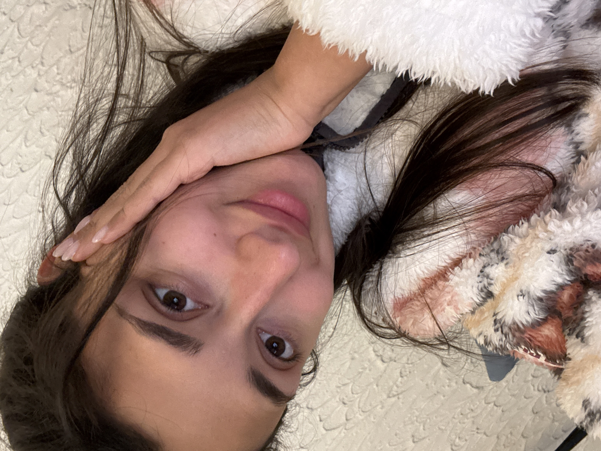 A close-up, angled image depicts a female individual, her head and upper torso positioned diagonally within the frame. She has long, dark brown hair, light brown eyes, and dark eyebrows, looking directly forward with a subtle smile. Her left hand, featuring light-colored manicured fingernails, rests against her left cheek. She is dressed in a white, high-pile, textured garment, possibly fleece or sherpa, which partially covers a darker layer visible at the neck. A second multi-colored, patterned, and similarly textured fabric, appearing as a blanket or soft furnishing, is visible in the lower left portion of the image. The background consists of a light-toned wall with an irregular, raised texture. The location is the city of Sutomore, Montenegro.