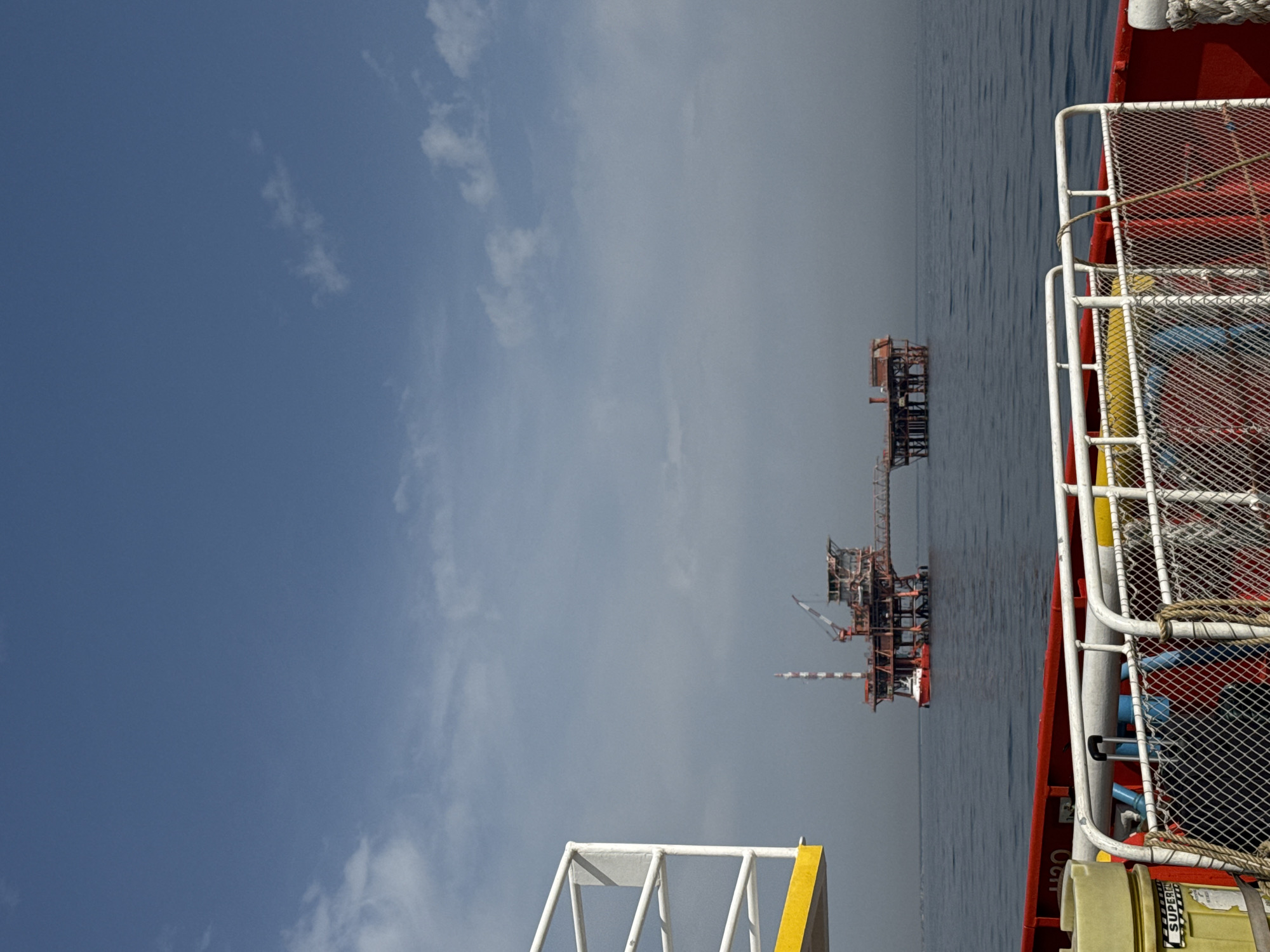 An offshore oil rig stands in the distance on a calm sea under a hazy blue sky. In the foreground, the railing of a boat or dock is visible, with a yellow structure on the left and a red structure on the right. The oil rig, predominantly red and brown, is a complex structure with multiple levels and a crane. A smaller red vessel is docked at the base of the rig. The scene is likely located in the Adriatic Sea, near the coast of Marina di Ravenna, Italy, a region known for its offshore oil and gas platforms. There are no people visible in the image.