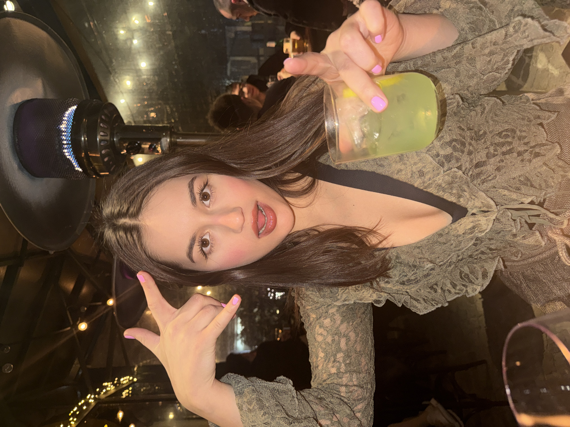 A young woman is depicted in a close-up, low-light photograph, set within an outdoor or semi-enclosed bar or restaurant in Sofia, Bulgaria. She faces the viewer directly, her mouth slightly agape with the tongue visible, and her brown eyes open. Her long, dark brown hair is parted centrally. She exhibits light pink blush on her cheeks and light pink lipstick. Her fingernails are painted a consistent light pink. She wears a khaki-green, lace-patterned top featuring ruffled trim, with a black undergarment strap visible at her neckline. Her left hand is raised, positioned to the left of her head, forming a 'rock-on' gesture with extended index and pinky fingers, while her thumb, middle, and ring fingers are folded. Her right hand holds a clear glass containing a light green liquid, ice cubes, and a lemon wedge; her index finger points forward from the glass, extending beyond the rim.
The background is dimly lit, featuring warm, spherical string lights, some of which are reflected on what appears to be a glass partition. A dark metal outdoor patio heater, emitting a blue flame from its mesh top, is suspended above and to the right of the woman. Blurred figures of other patrons are visible behind her, including a man with dark hair holding a beer glass to the right. A partial view of a wine glass occupies the lower left foreground. The precise establishment name or address within Sofia, Bulgaria, cannot be determined from the provided visual evidence.