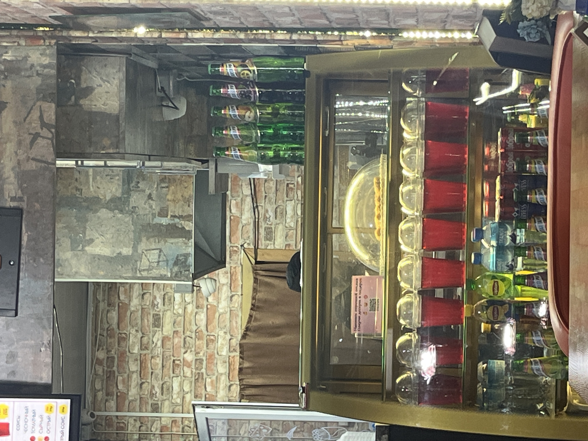 The image shows a shelf stocked with various drinks, some in bottles and others in red plastic cups. The bottles appear to be soft drinks and water, with labels visible. The red cups are filled with a red liquid and are stacked in rows. Behind the drinks, there is a display case with a golden frame. In the background, there are brick walls, suggesting an indoor setting such as a cafe or bar. The lighting appears to be artificial, with some lights reflecting off the bottles and cups. There are no people clearly visible in the image, but there might be a partial silhouette of a person behind the display case. Some text is visible on some of the bottles and on a small pink sign in the display case, but it is too blurry to read. The overall impression is of a commercial establishment selling beverages. The orientation of the image suggests it was taken with the camera held sideways.