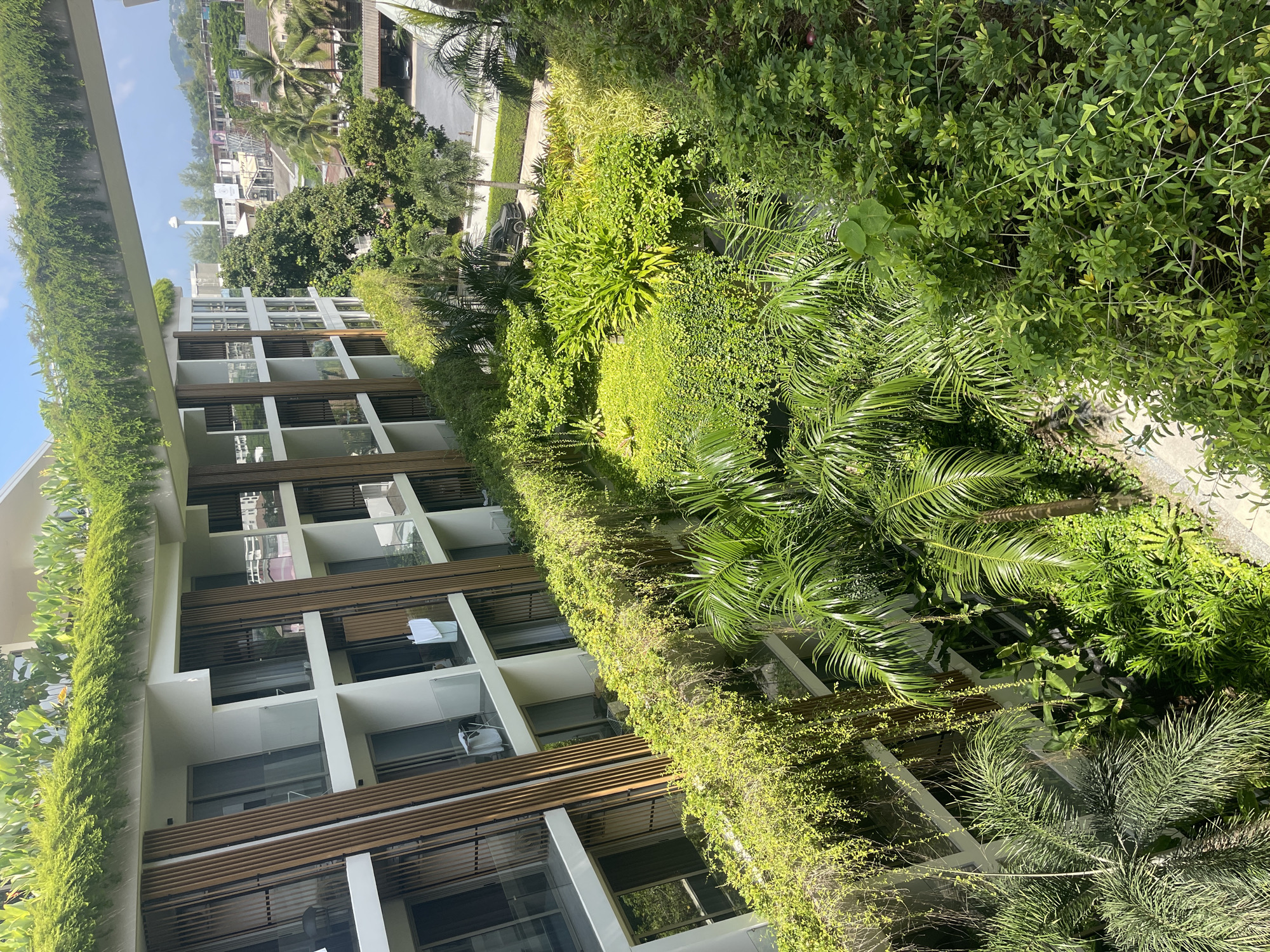 The image shows a modern building with balconies covered in lush green vegetation, likely in the city of Choeng Thale, Thailand. The building appears to be a hotel or condominium, with multiple levels of rooms, each featuring glass doors leading to balconies. Vertical wooden louvers are interspersed between the balconies, providing shade and architectural detail.
A significant feature of the scene is the extensive greenery cascading over the balconies and forming a dense garden on lower levels. This includes various types of plants, such as palm trees, vines, and other tropical foliage, creating a vibrant and natural aesthetic. The dense vegetation suggests a tropical climate, consistent with the reported location.
In the background, other buildings and palm trees are visible, indicating an urban or resort environment. A water tower can be seen in the distance, along with a clear sky. A black car is parked on a road in the middle ground. There are no people clearly visible in the image, but laundry can be seen hanging on one of the balconies, suggesting occupancy. The overall impression is one of a well-maintained, nature-integrated architectural design.