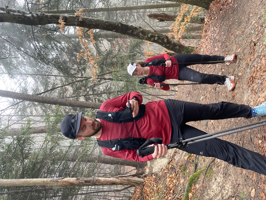 Two individuals are depicted on a sloped, leaf-strewn dirt trail within a forested area, consistent with the terrain found near Sallanches, France, under overcast or misty atmospheric conditions.
In the foreground, a male with a beard wears a black baseball cap (with a "BUFF" logo), a red long-sleeved athletic shirt (with "SALOMON" visible on the left sleeve), a black Salomon hydration vest, black athletic pants (with "KALENJI" visible on the left thigh), and light blue, orange, and white Salomon trail running shoes. He holds a black smartphone in his right hand, looking down at its screen, and a pair of collapsed black and green hiking poles in his left hand.
Behind him and slightly to his right, a female wears a white baseball cap (with a "BUFF" logo), a red long-sleeved athletic shirt, a black hydration vest similar to the male's, black athletic pants, and white and orange trail running shoes. She holds a pair of black hiking poles, with their tips on the ground, one in each hand. A dark-colored sports watch is visible on her left wrist. She is looking to her right.
The environment consists of a densely wooded area with a mix of conifer and deciduous trees. The ground is covered with reddish-brown fallen leaves. The background shows trees obscured by a general haze or mist, indicating cool or damp weather.