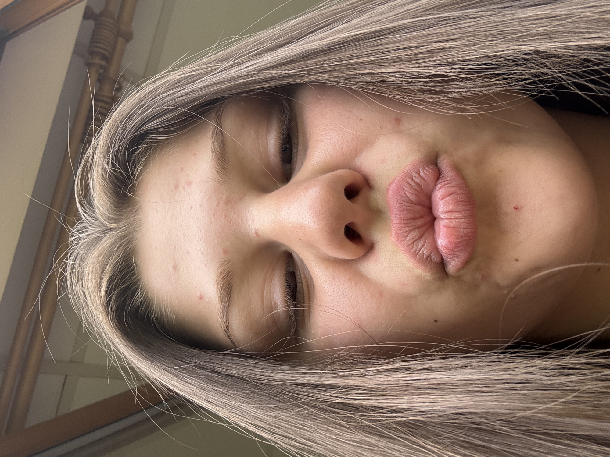 A close-up, low-angle, indoor shot shows a young person with light brown hair making a kissing face. Their eyes are partially closed and their lips are puckered. The person's skin has a few blemishes. Their hair is parted in the middle and falls around their face. The background is out of focus and appears to be a wall with some wooden trim.