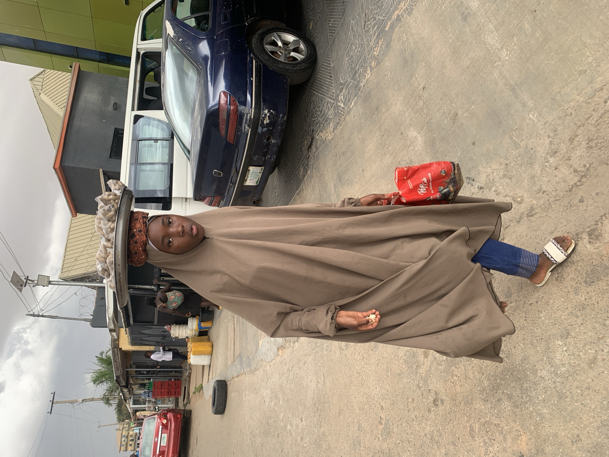 A young woman wearing a dark brown hijab and blue pants stands on a paved street. She is carrying a red bag in her left hand and what appears to be small white food items in her right. A tray is balanced on her head, containing what looks like bundled items. Behind her are parked vehicles, a blue sedan and a white van. In the background are buildings and other people. A red car is parked further down the street along with a black tire. The setting appears to be an urban or commercial area. The location is Jos, Nigeria.