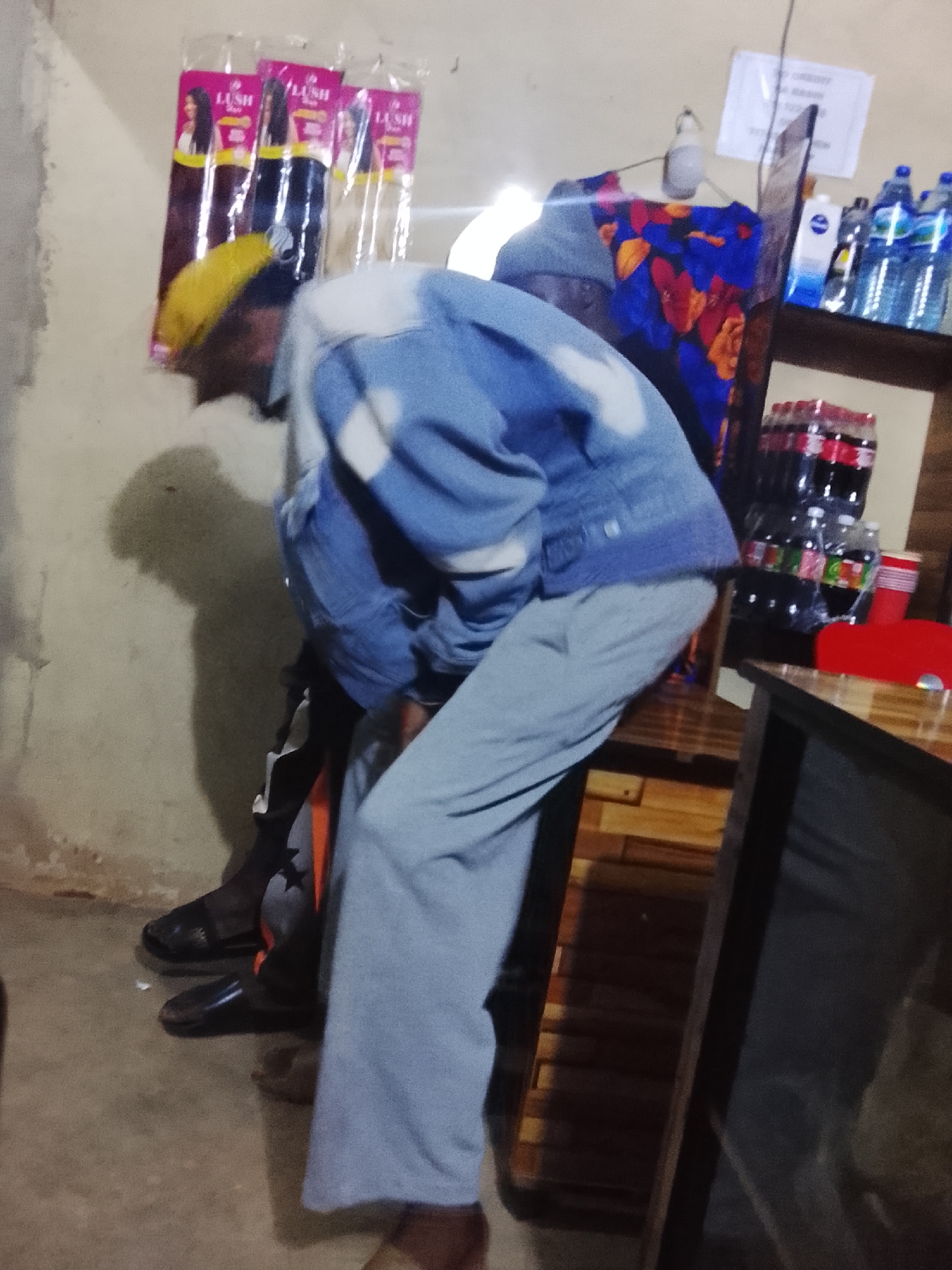 The image shows a person wearing a blue denim jacket with white accents and light blue pants, with a yellow cap on their head. They appear to be hunched over, possibly engaged in some activity. The setting is indoors, with a wall that has some texture and discoloration. In the background, there are several items displayed on shelves, including bottles of water and what appear to be Coca-Cola bottles. There are also packages of hair extensions hanging on the wall. A sign is visible on the wall with some text that is not clearly legible. The lighting in the room appears to be artificial, suggesting it could be daytime or nighttime. The overall impression is that of a small shop or store in Durumi, Nigeria. The floor is concrete, and there is a shadow cast on the wall behind the person. Another person's feet are visible in the lower left of the frame, wearing black sandals. The scene seems casual and everyday.