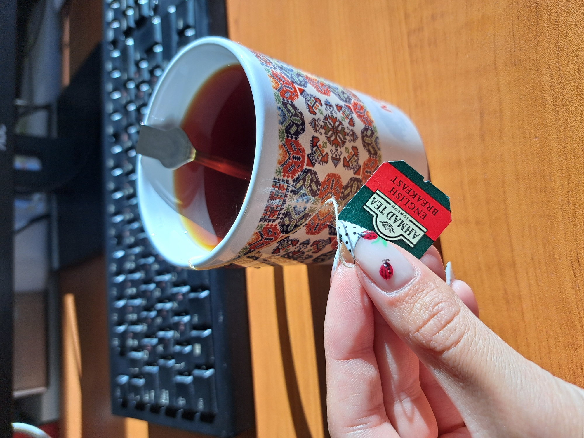 A close-up shot shows a hand holding a tea bag over a patterned mug filled with dark liquid. A spoon is inside the mug. The tea bag's label reads "AHMAD TEA ENGLISH BREAKFAST". The hand features manicured nails with a ladybug design. In the background, a black computer keyboard is visible on a wood-like surface. The location appears to be Unknown, Unknown.
