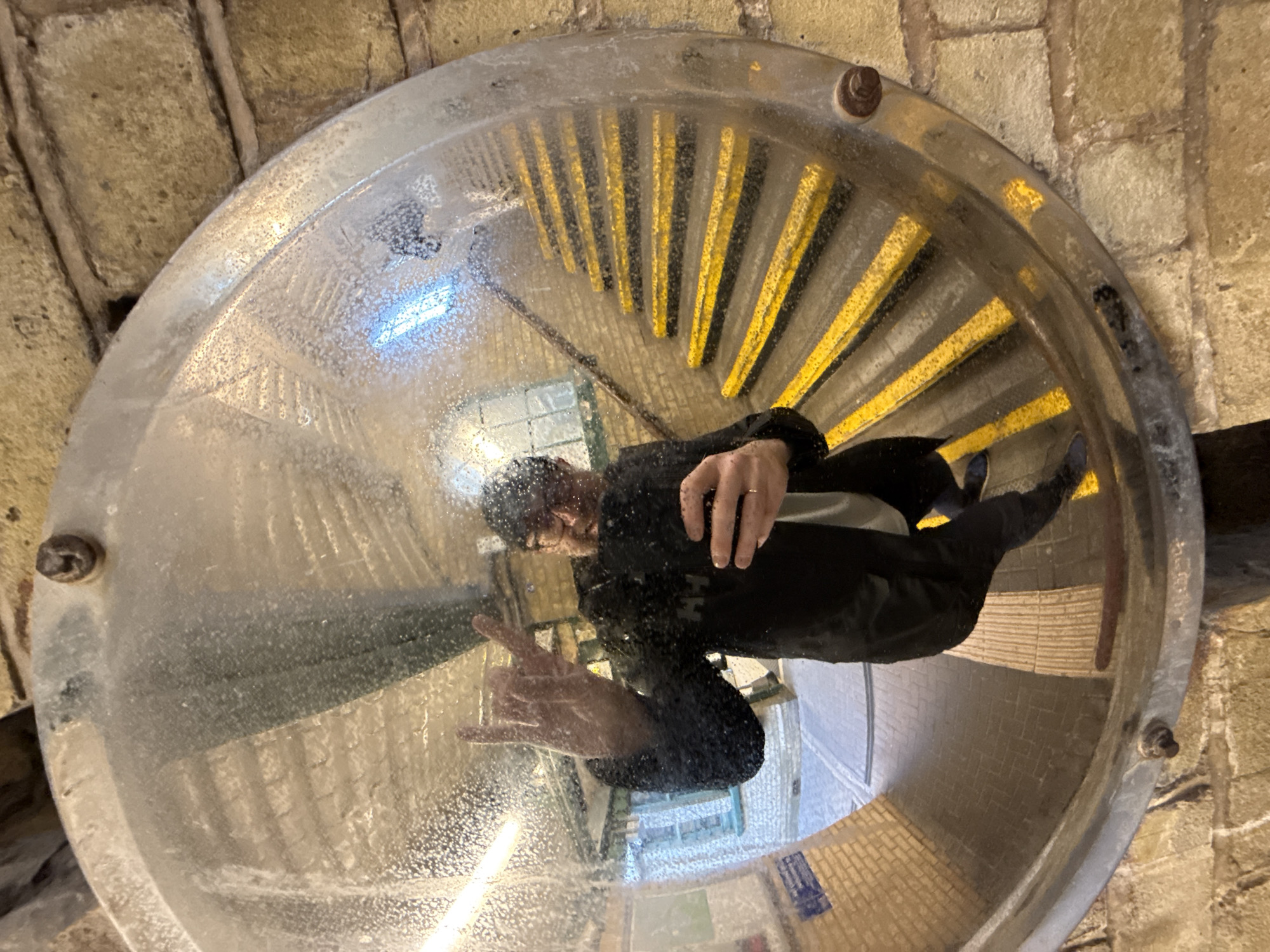 The image shows a reflection in a convex mirror mounted on a brick wall. The reflection captures a person in a black coat and glasses, giving a "rock-on" hand gesture. The reflection also shows a set of stairs with yellow and black striped steps. The presence of the stairs and the brick wall suggest the location is likely a London Underground station.